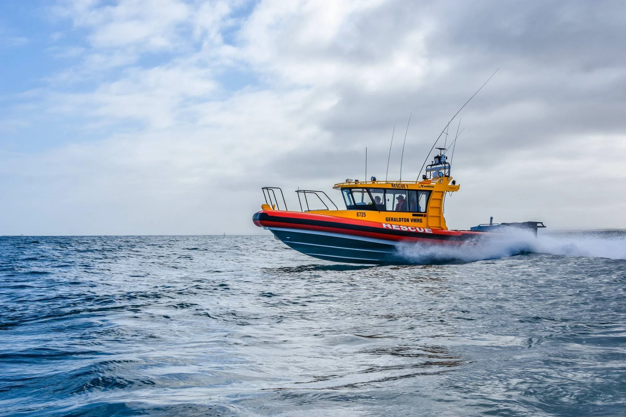 A yellow rescue boat moving swiftly across the water with waves splashing behind it, under a cloudy sky.