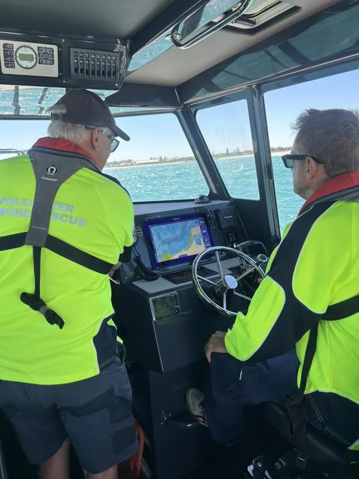 Two people inside a boat's cockpit, both wearing bright yellow life jackets with 'Volunteer Marine Rescue' on the back, looking at a GPS navigation screen while steering the boat on a body of water.