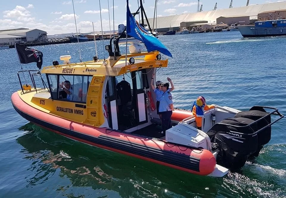 A rescue boat with officials aboard on a sunny day in a marina with other boats and docks in the background.