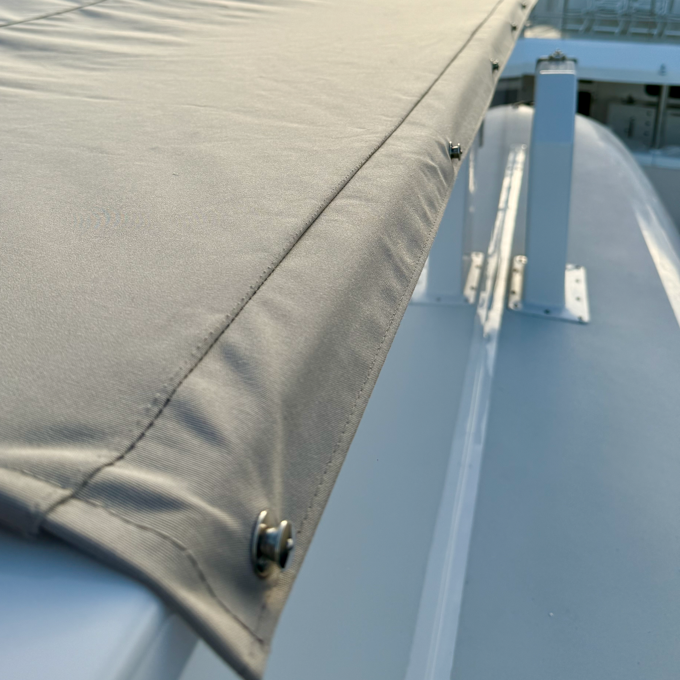 Close-up of a beige fabric canopy on a boat, secured with metal fasteners, with the boat's white surface and railings visible in the background.