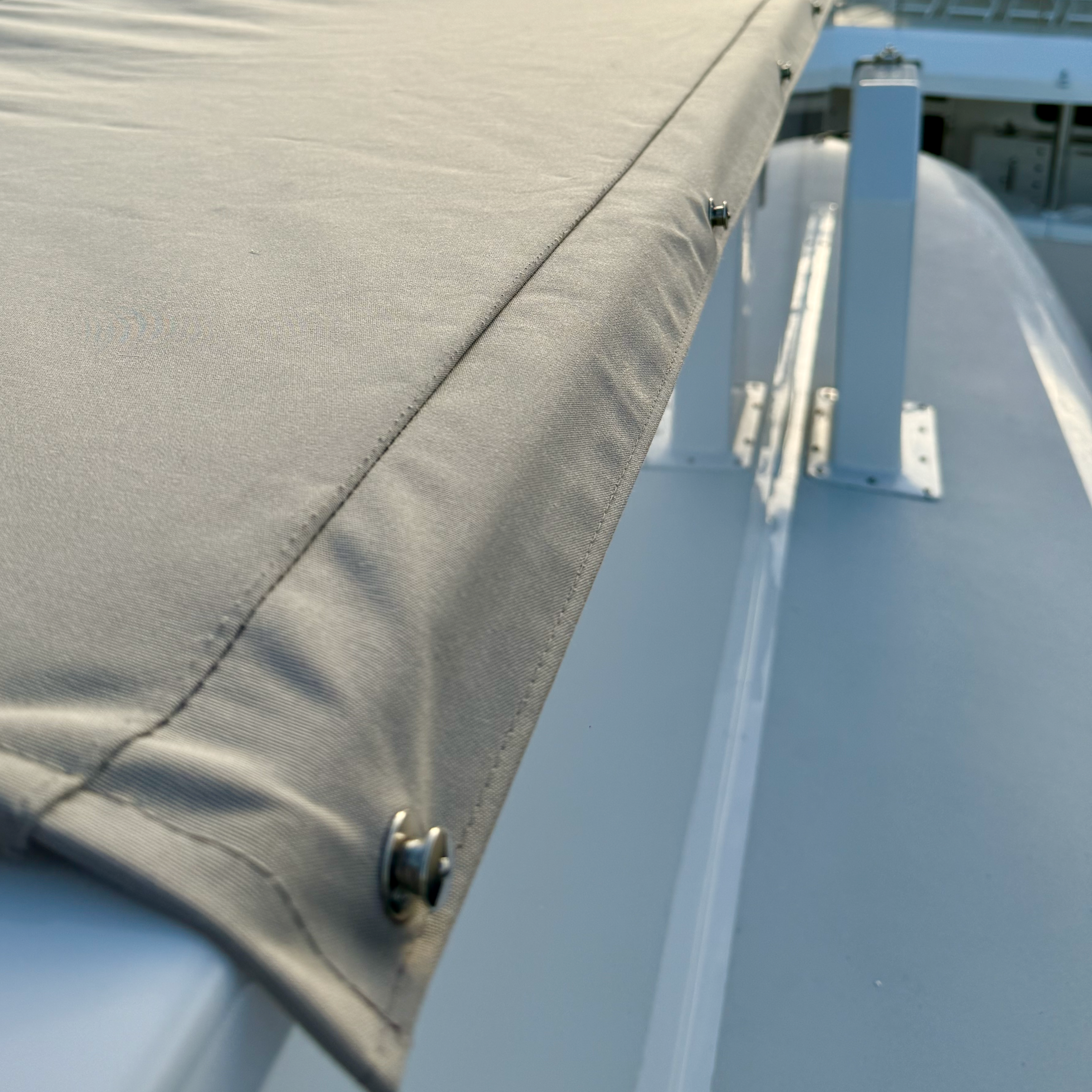 Close-up of a metallic cover with a beige fabric edge and rivets, mounted on a white surface, possibly part of a boat or an industrial object.