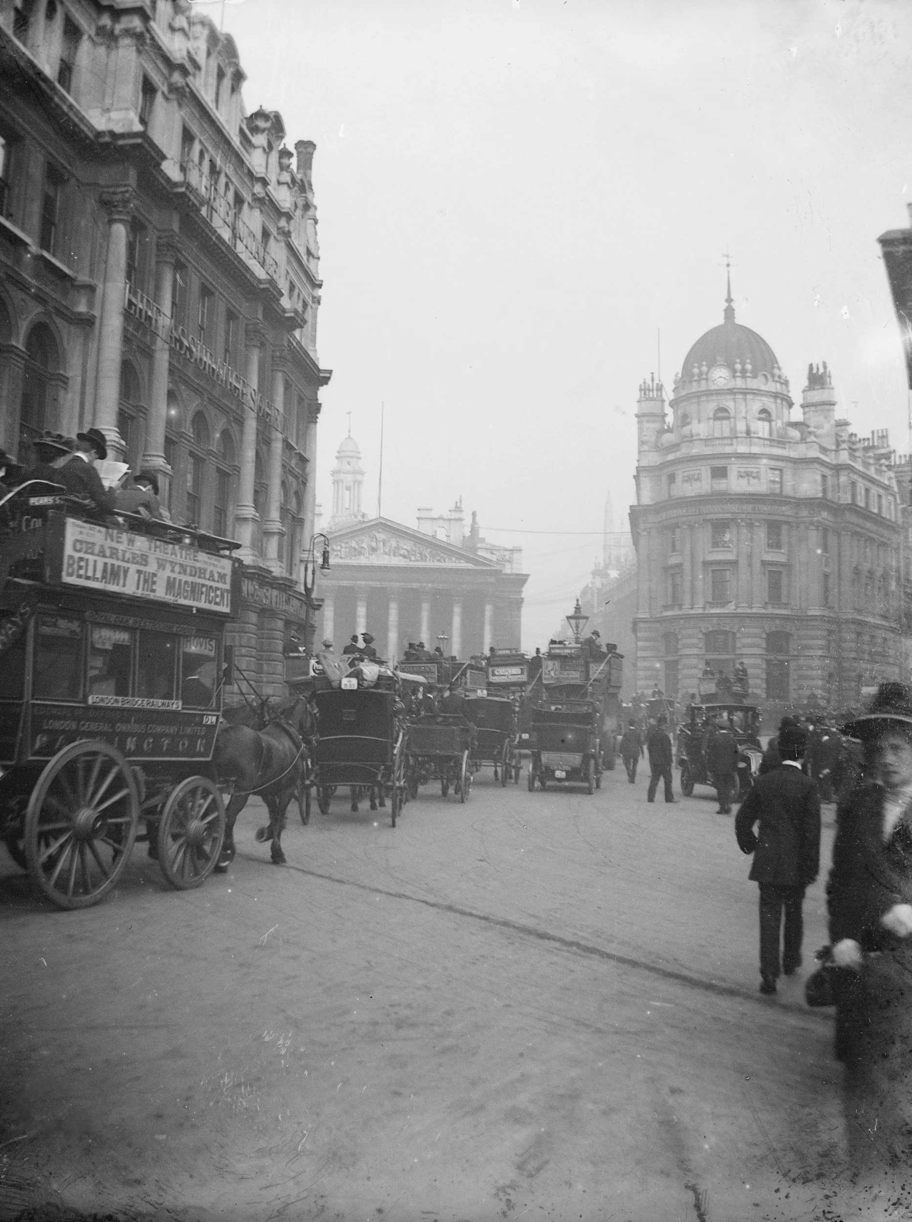 Historical street scene with horse-drawn carriages, pedestrians, and early 20th-century buildings with signs, including a theatre marquee advertising Charles Wyndham and Bellamy the Magnificent.