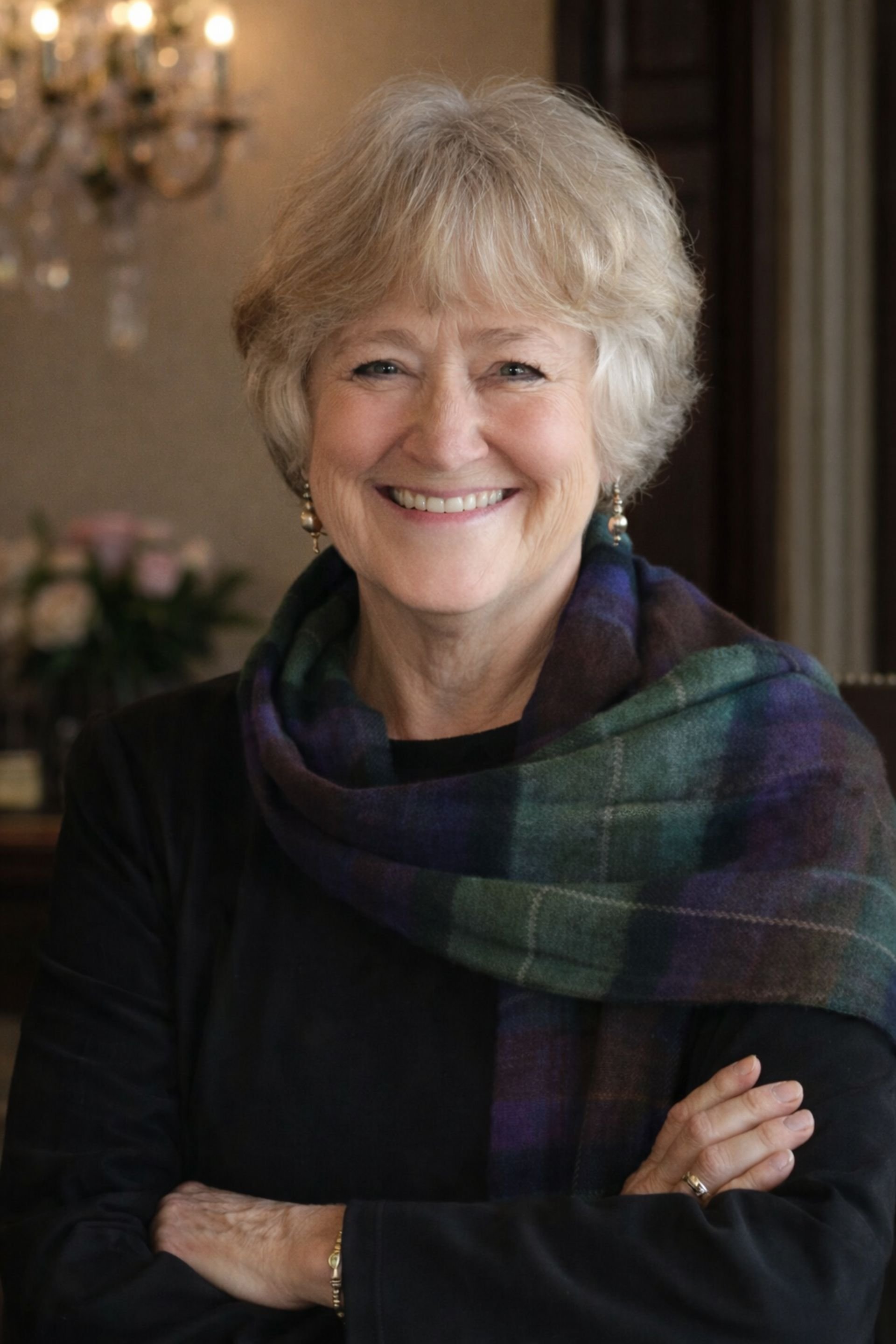 Historical time travel fiction author Helen Pilz, wearing earrings, a dark jacket, and a multicolored plaid scarf, standing indoors with a chandelier and blurred floral arrangement in the background.