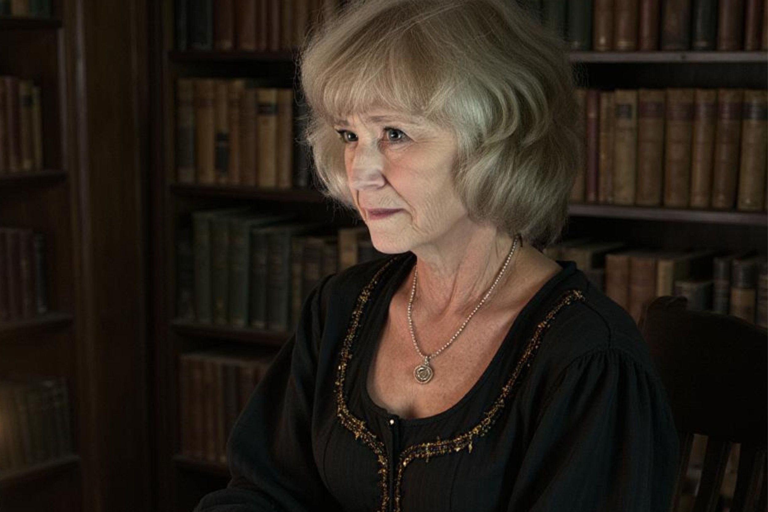 Historical time travel fiction author Helen Pilz, in a black embroidered blouse, sits in front of a bookshelf filled with leather-bound books.