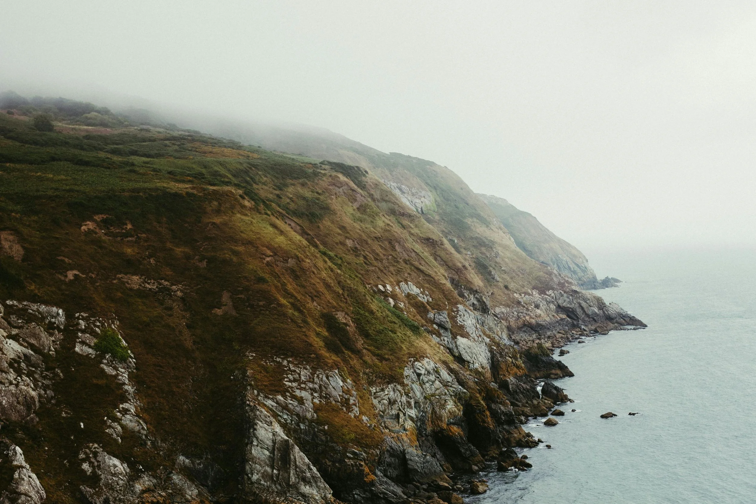 Coastal cliffs with green vegetation and rocks, partially covered in fog, meeting the ocean.