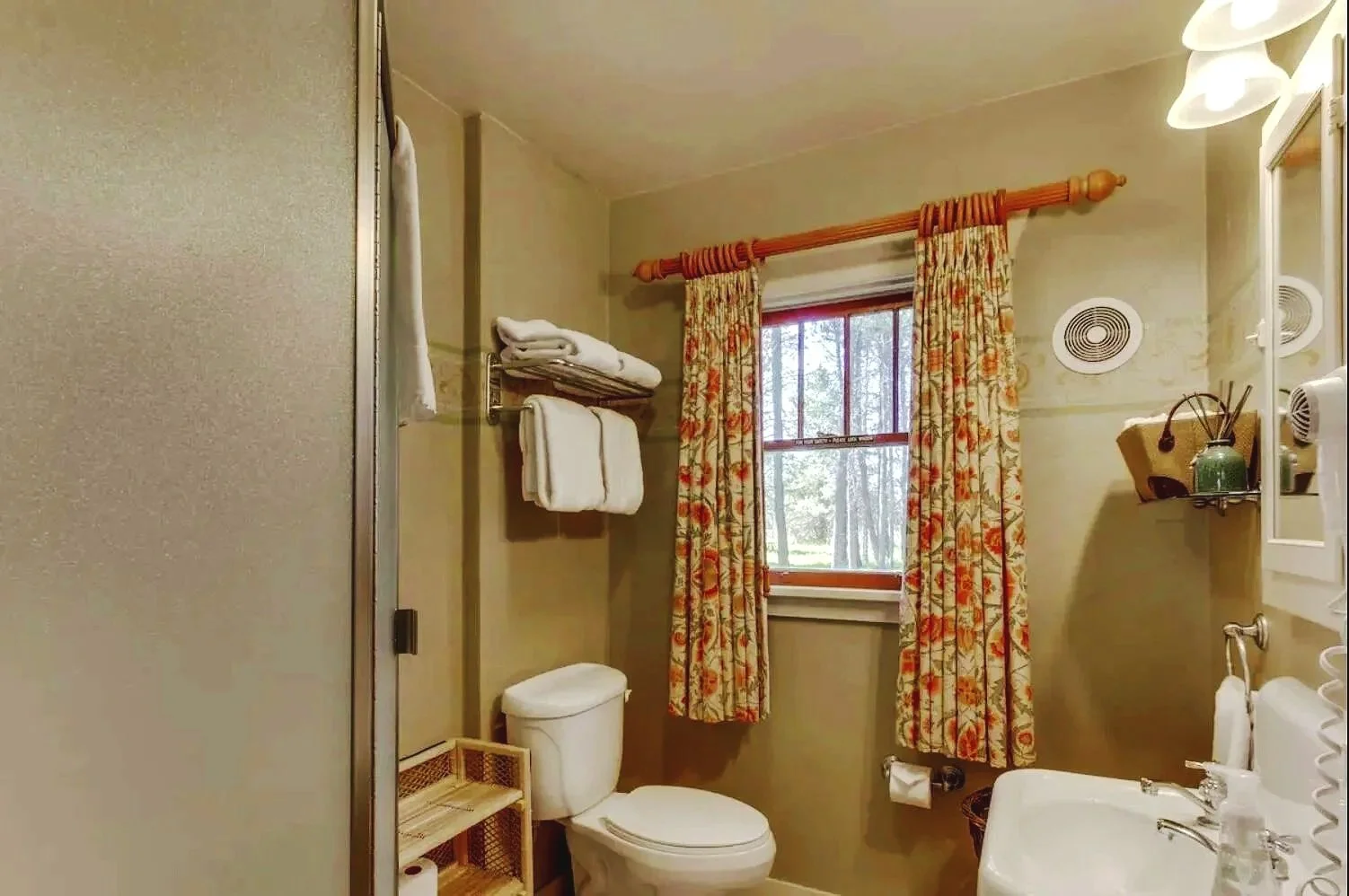 A cozy bathroom with a window showing trees outside, decorated with floral curtains, white towels on a metal rack, a white toilet, a small wooden shelf, and a sink with toiletries. The room has beige walls and a ceiling light fixture.
