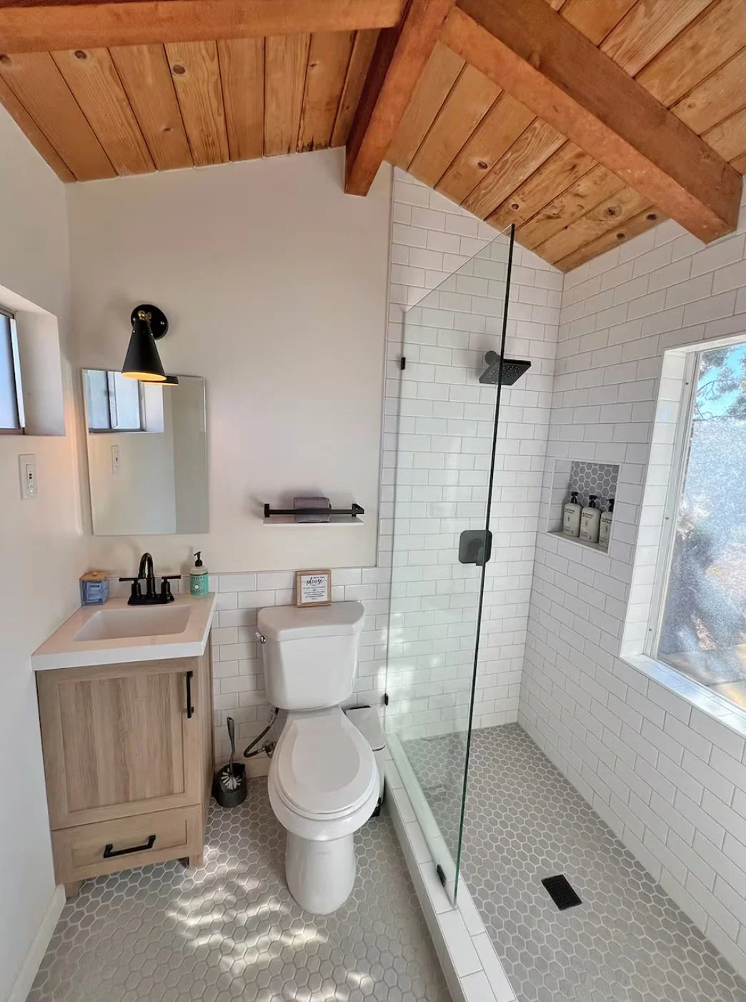 A modern bathroom with a wooden ceiling, white tile walls, a glass-enclosed shower, a small wooden vanity with a sink, a black wall-mounted lamp, a mirror, and a window with natural light.