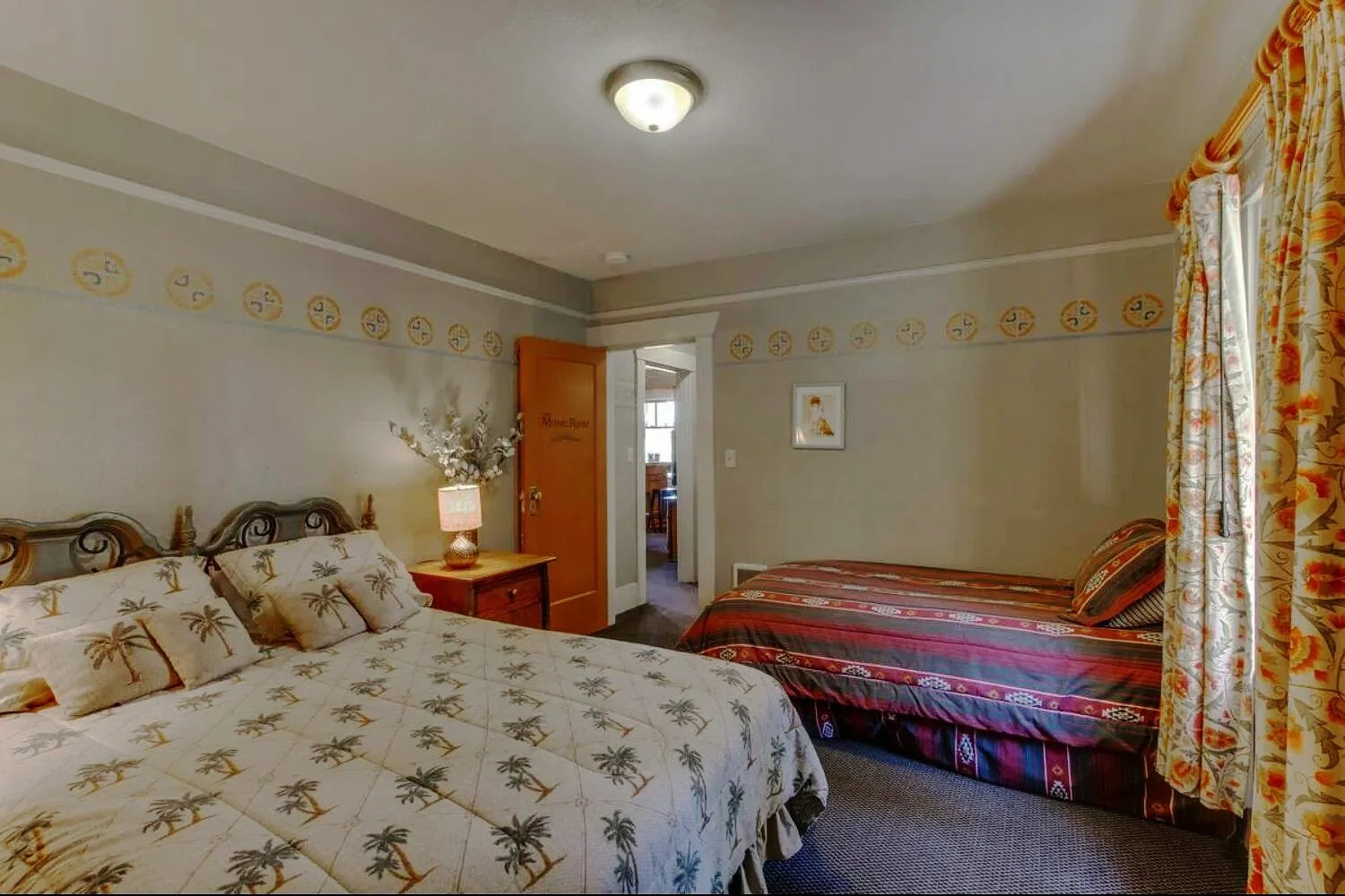 A room with two beds, one with a white palm tree patterned bedspread and pillows, and another with a striped bedspread and pillows. There is a nightstand with a lamp and decorative flowers, and a window with floral curtains. The walls have a circular border with a yellow and blue pattern, and a framed picture hangs on the wall.