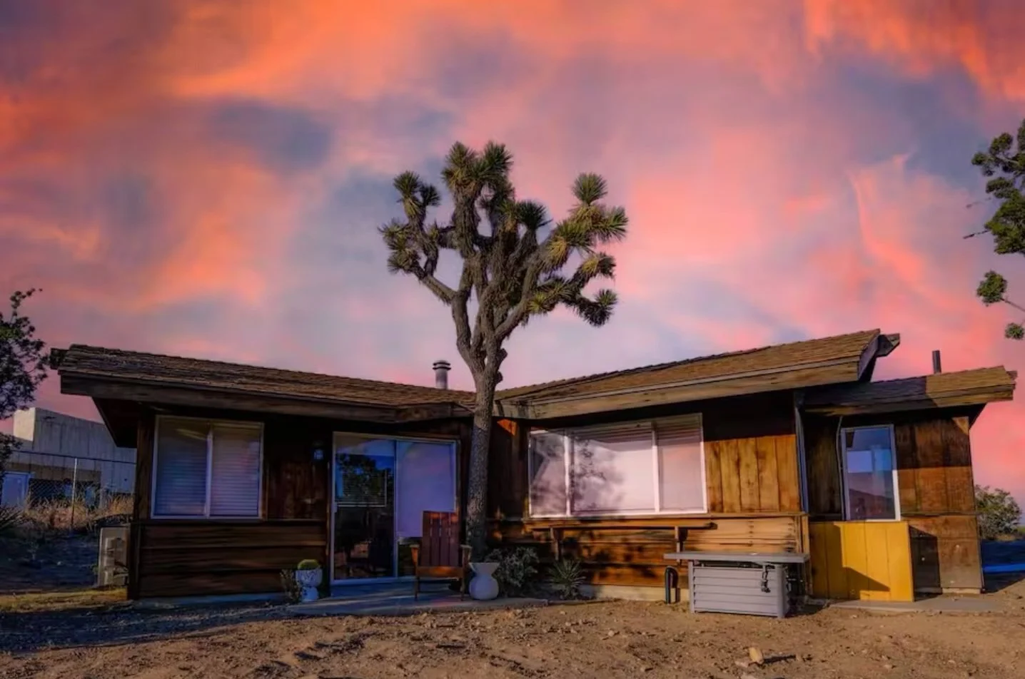 A single-story house with a unique, angular roof design, large windows, and wooden exterior siding. There is a Joshua tree, growing in front of the house, set against a colorful sunset sky with pink and orange clouds.