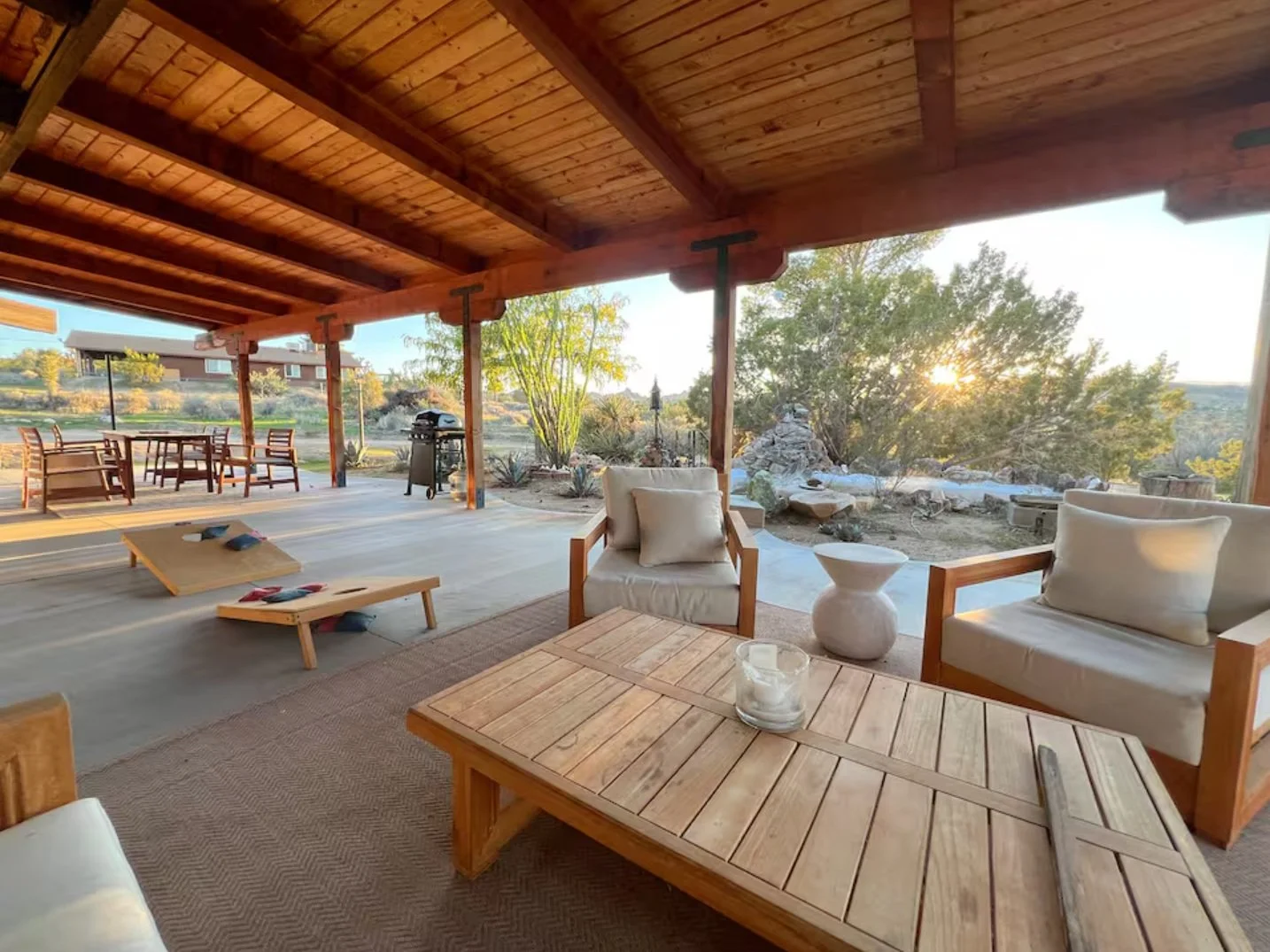 Sunset view from a spacious outdoor patio with wooden furniture, including a coffee table, chairs, and a sofa, overlooking desert landscape with trees, rocks, and a fire pit.