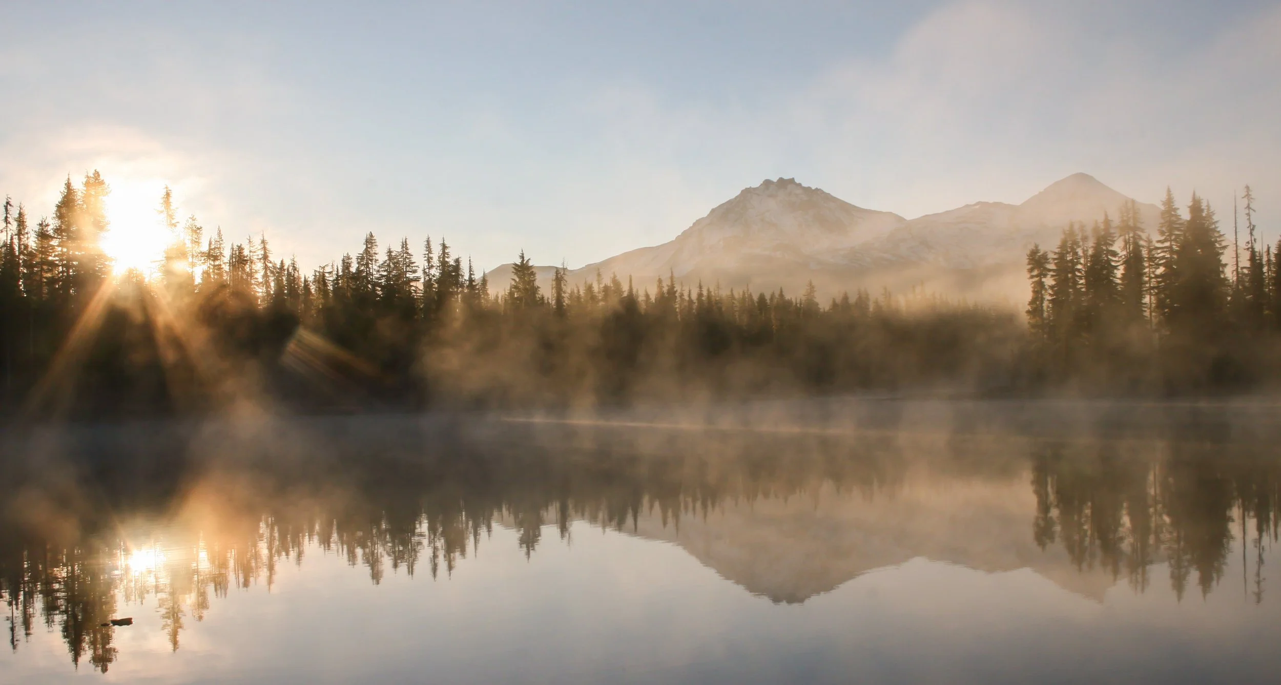 Sunrise over a mountain range reflected in a calm lake, with a forest of pine trees and mist rising from the water.