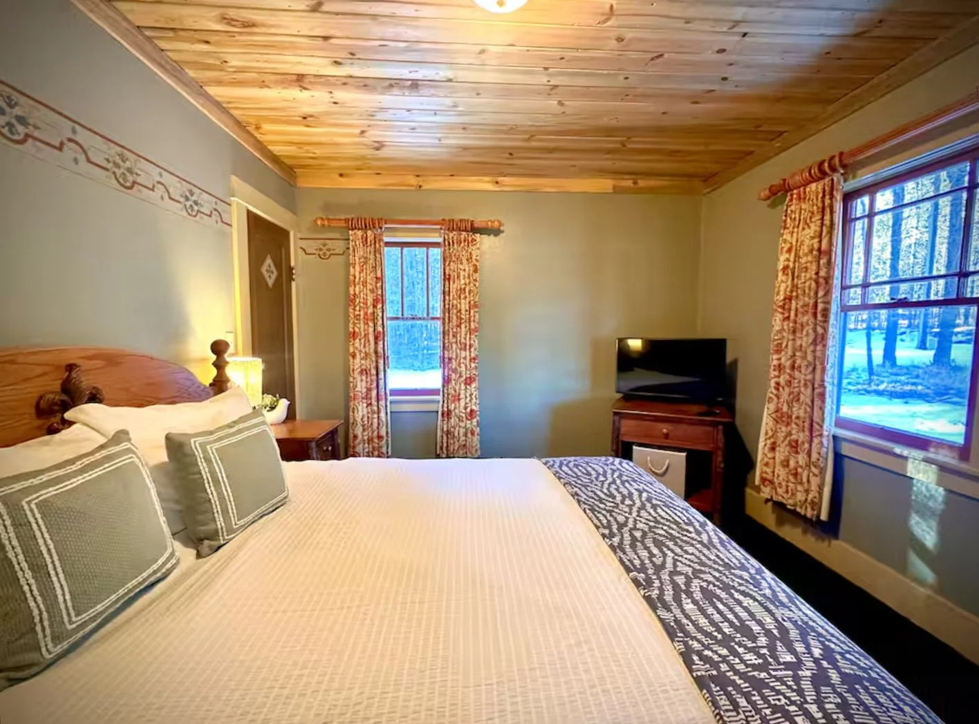 Cozy bedroom with wooden ceiling, light-colored walls, a large bed with multiple pillows, bedside table with lamp, TV on wooden stand, and windows with floral curtains overlooking a snowy outdoor scene.