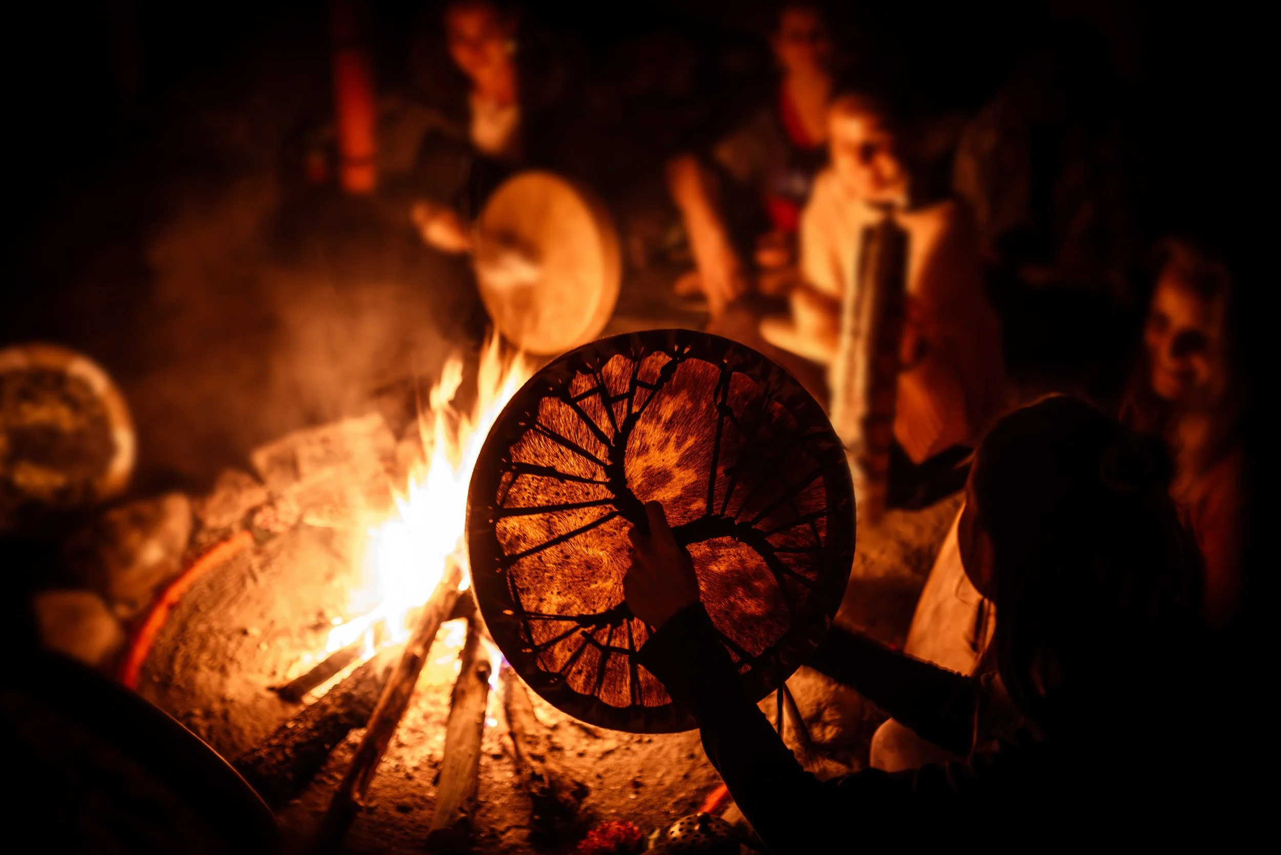 Person holding a hand drum near a fire pit with burning logs.