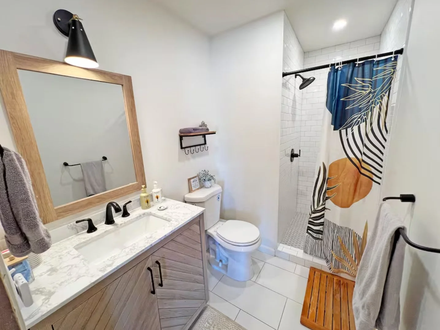 Bathroom with a vanity, mirror, towels, toilet, shower area with patterned shower curtain, and wooden bench.