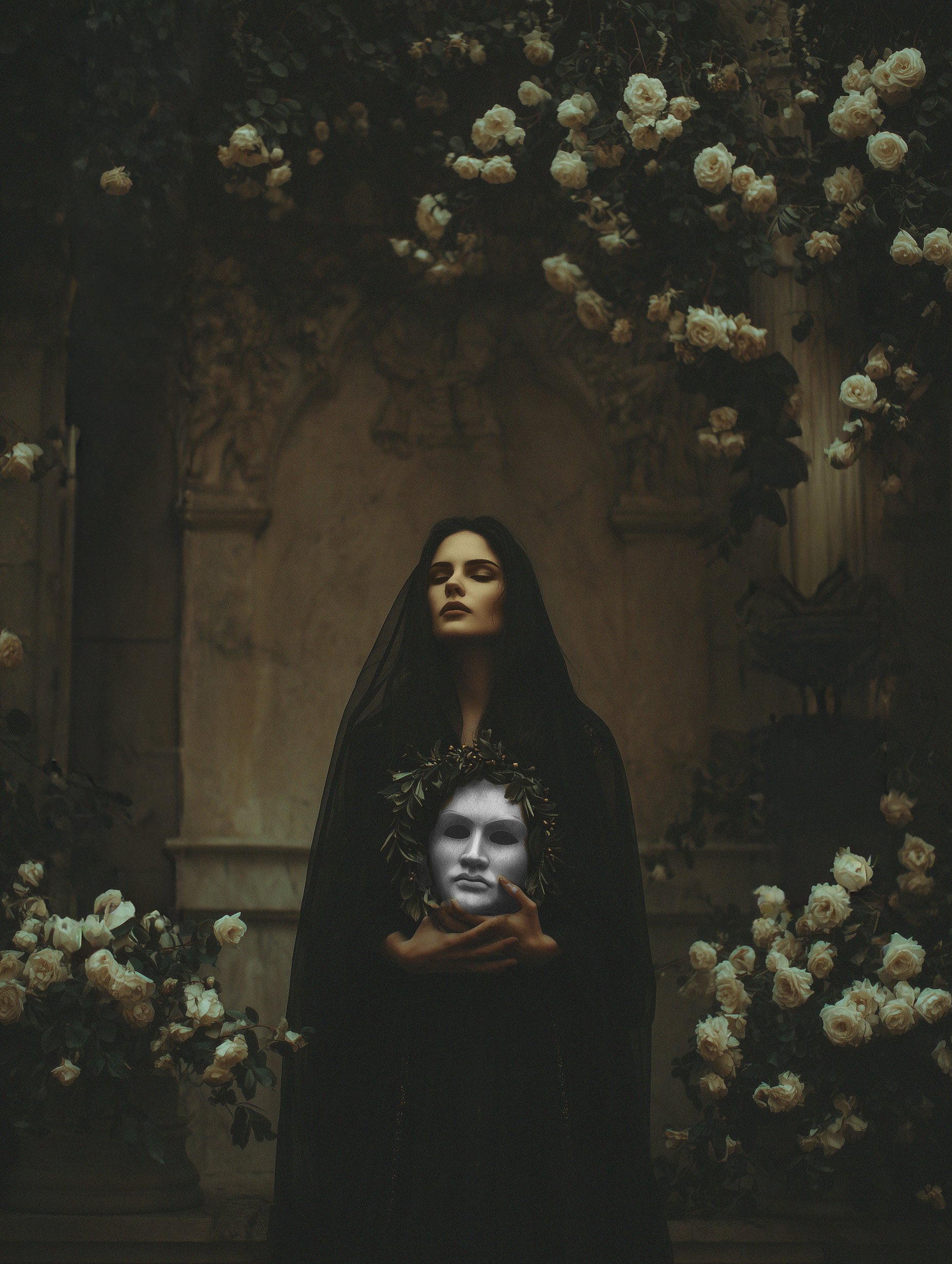 A woman dressed in black with dark hair and makeup, holding a white mask surrounded by white roses and dark foliage in a dimly lit setting.