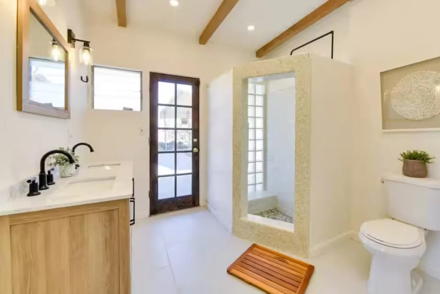 Modern bathroom with white walls, wooden accents, a shower enclosed with pebble tiles, a vanity with a black faucet, a toilet, a window, and a door leading outside.