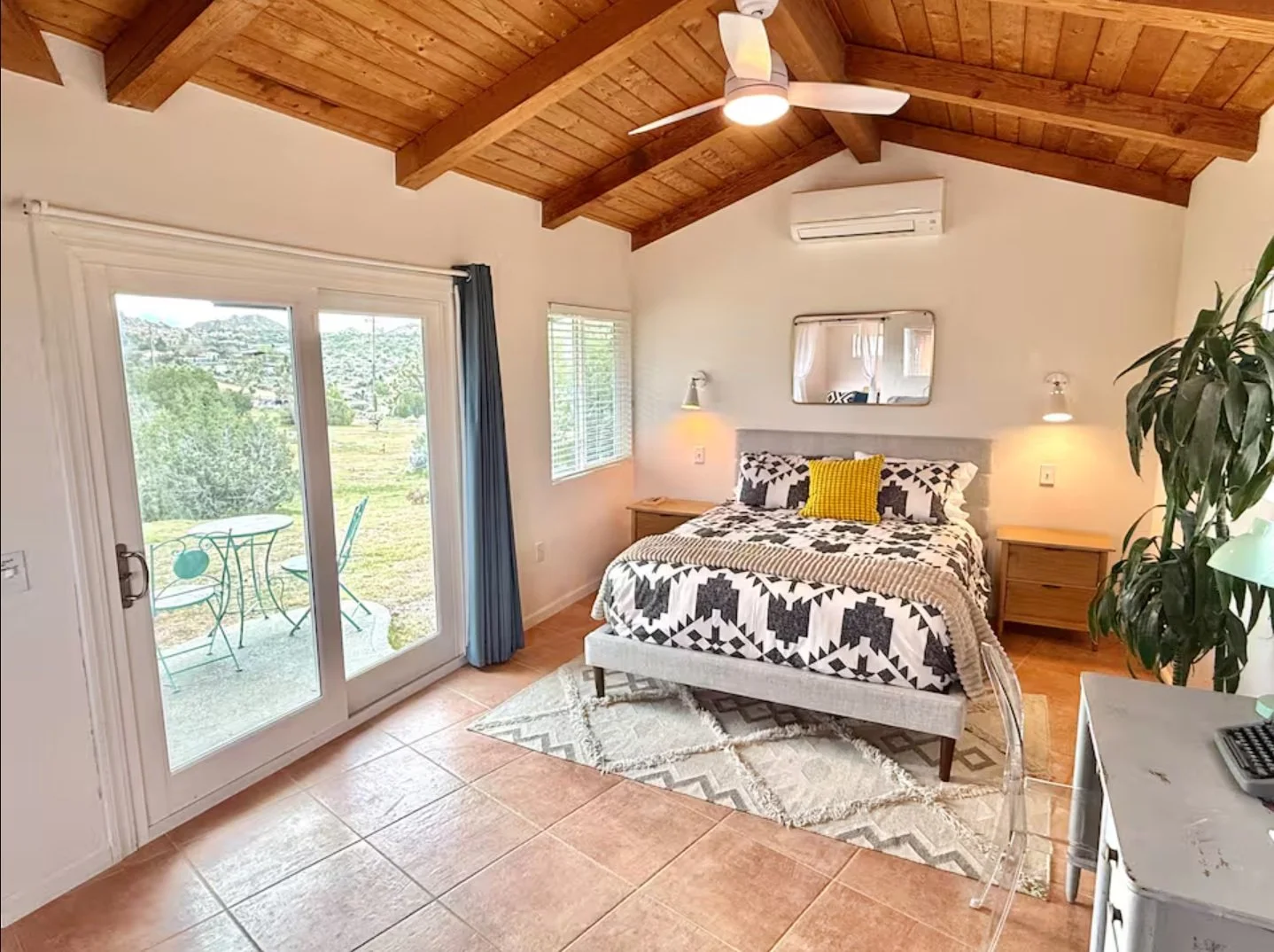 A bedroom with a wooden vaulted ceiling, a bed with black and white patterned bedding and yellow pillows, two wooden nightstands, a mirror above the bed, sliding glass doors to an outdoor patio, a small table and chairs outside, a large green plant, and a white dresser with a keyboard on it.