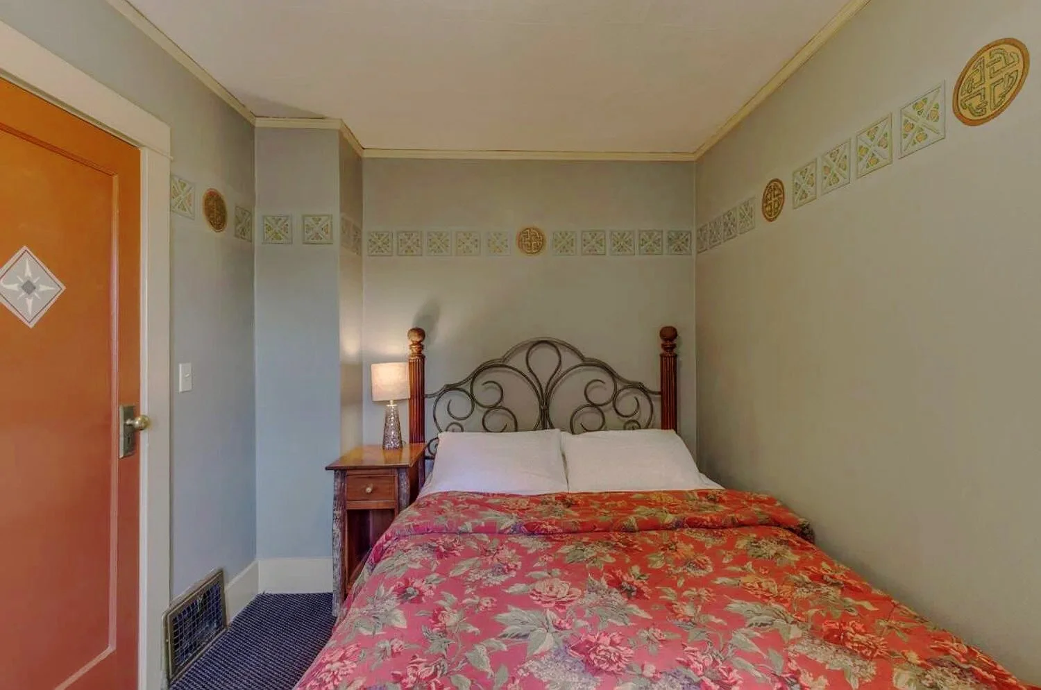 A bedroom with a bed covered in a floral pink and gold bedspread, white pillows, a wooden headboard, a nightstand with a lamp, and a wall decorated with a continuous border of patterned tiles and circular motifs.