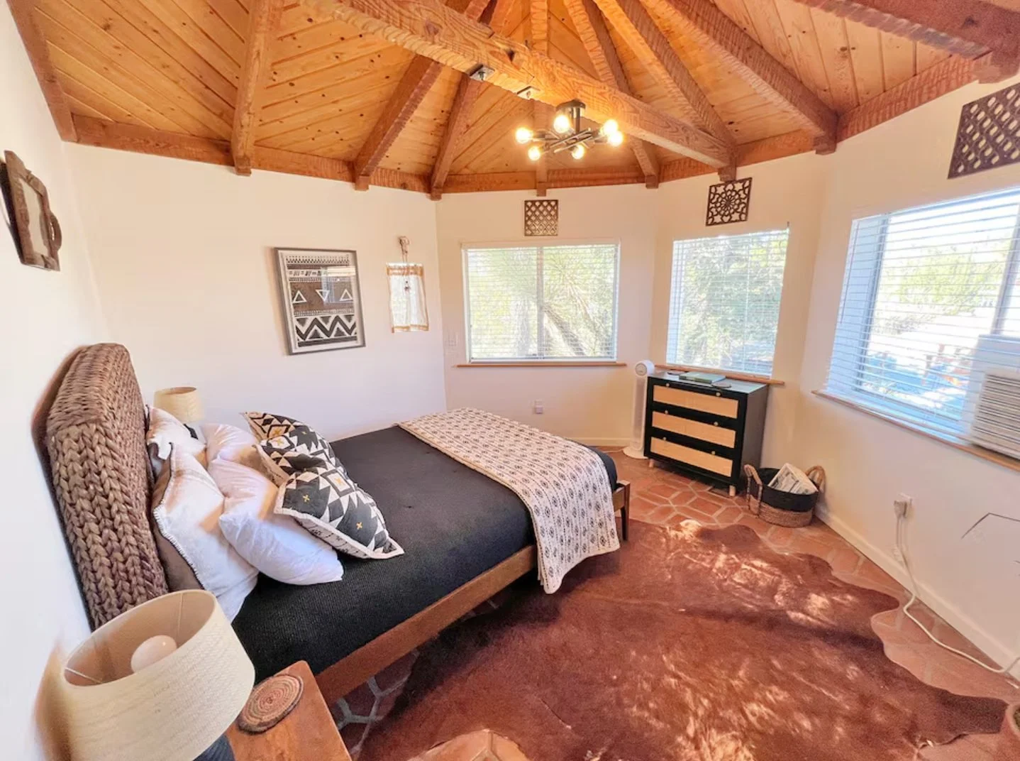 A cozy bedroom with a wooden vaulted ceiling, a bed with patterned pillows and a blanket, a nightstand with a lamp, three windows with blinds letting in natural light, a black dresser, and a cowhide rug on terracotta floor tiles.
