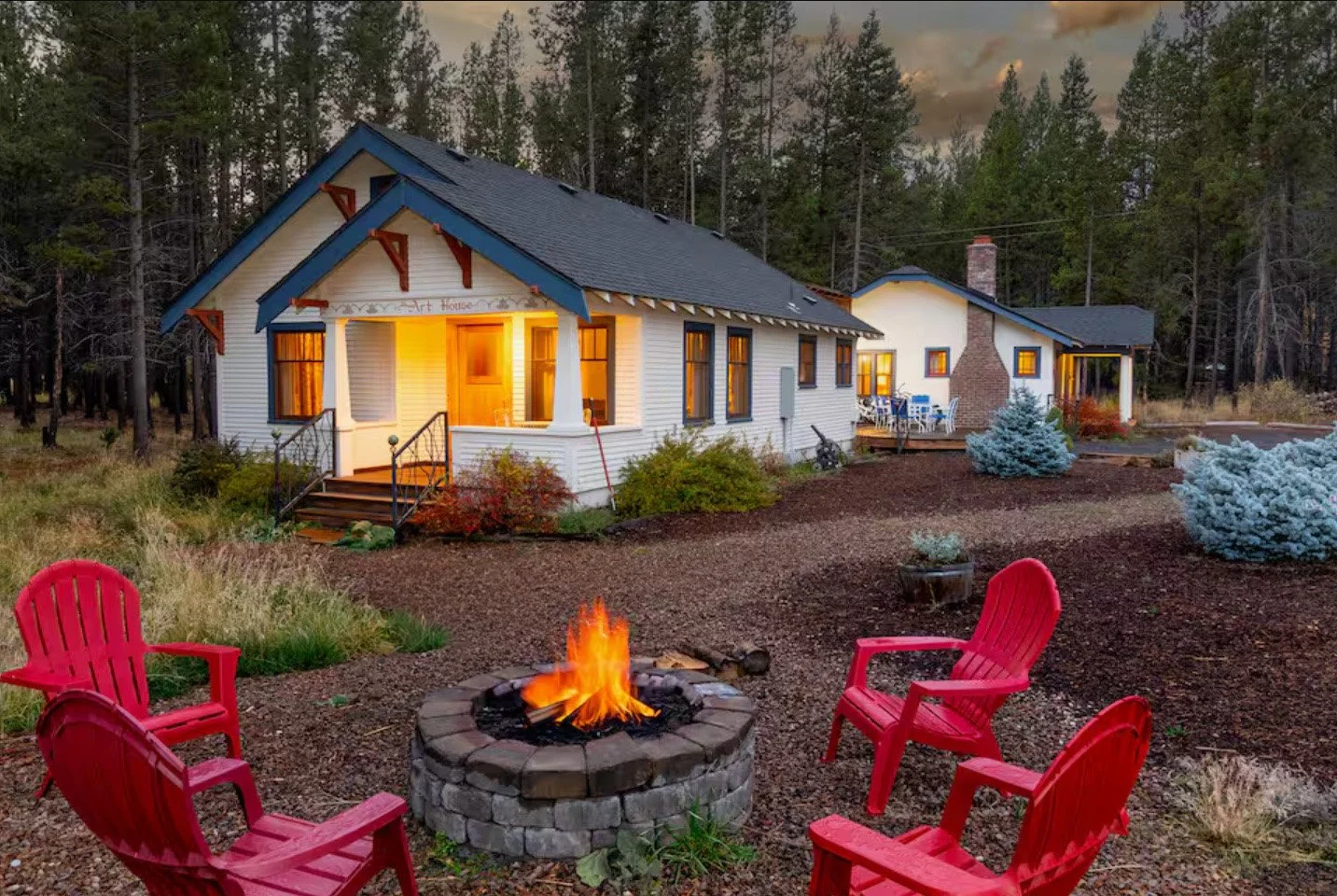 A cozy house with a lit fire in a firepit, surrounded by red chairs in a landscaped yard with trees in the background, during dusk.