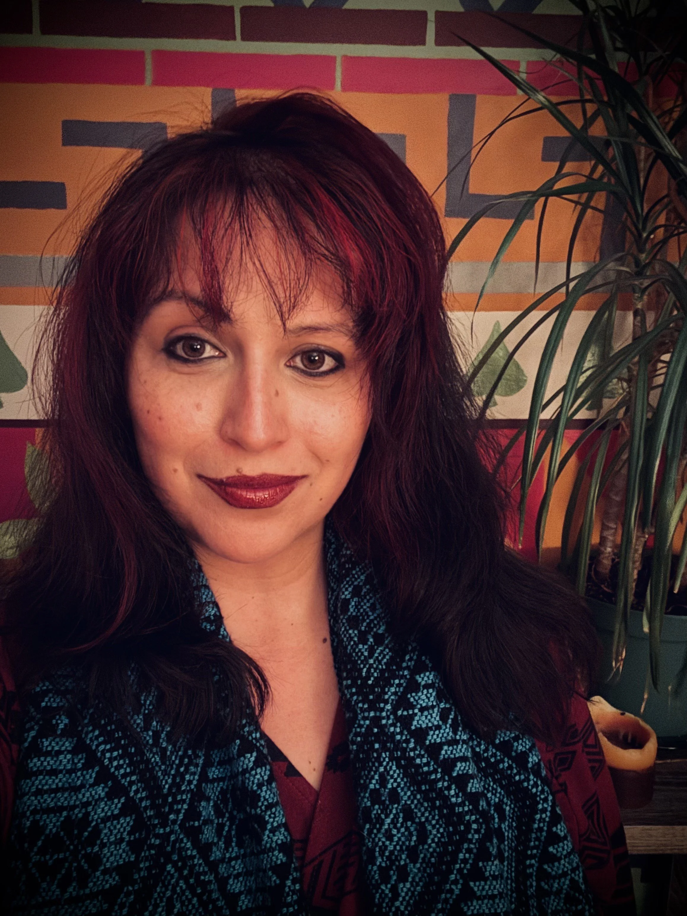 A woman with dark red hair, smiling with makeup, wearing a patterned top, with a background of a colorful painted wall and green plants.