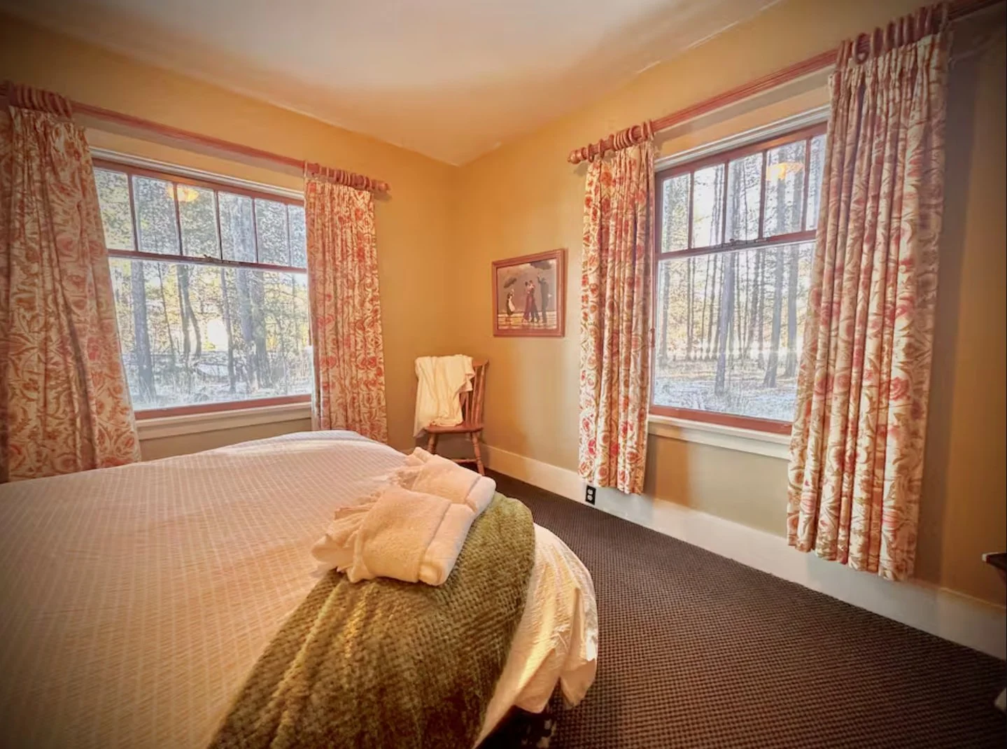 A cozy bedroom with two large windows with floral curtains, a bed with white linens and a green blanket, a wooden chair with towels on it, and a framed picture on the wall.