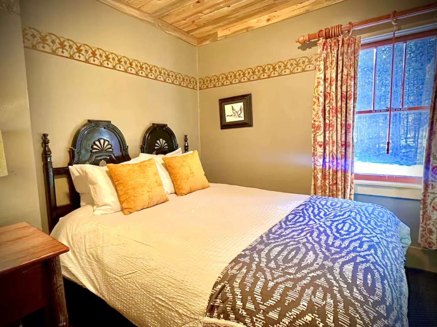 A cozy bedroom with a large bed featuring a dark wooden headboard, two yellow pillows, and a white quilt. To the right, a window with floral curtains showing a snowy outdoor scene. A small framed artwork hangs on the wall above the bed, and a wooden nightstand is partially visible on the left.
