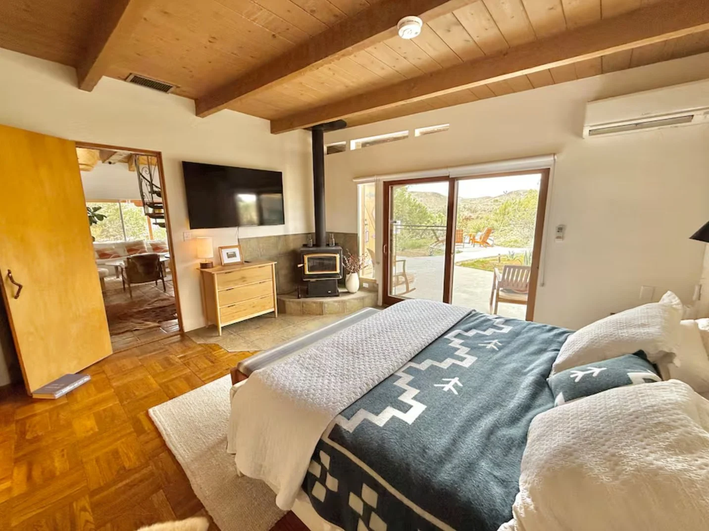 A cozy bedroom with a wooden ceiling, sliding glass door leading to an outdoor patio, a bed with white and blue bedding, a flat-screen TV mounted on the wall, a wood stove, and natural light.