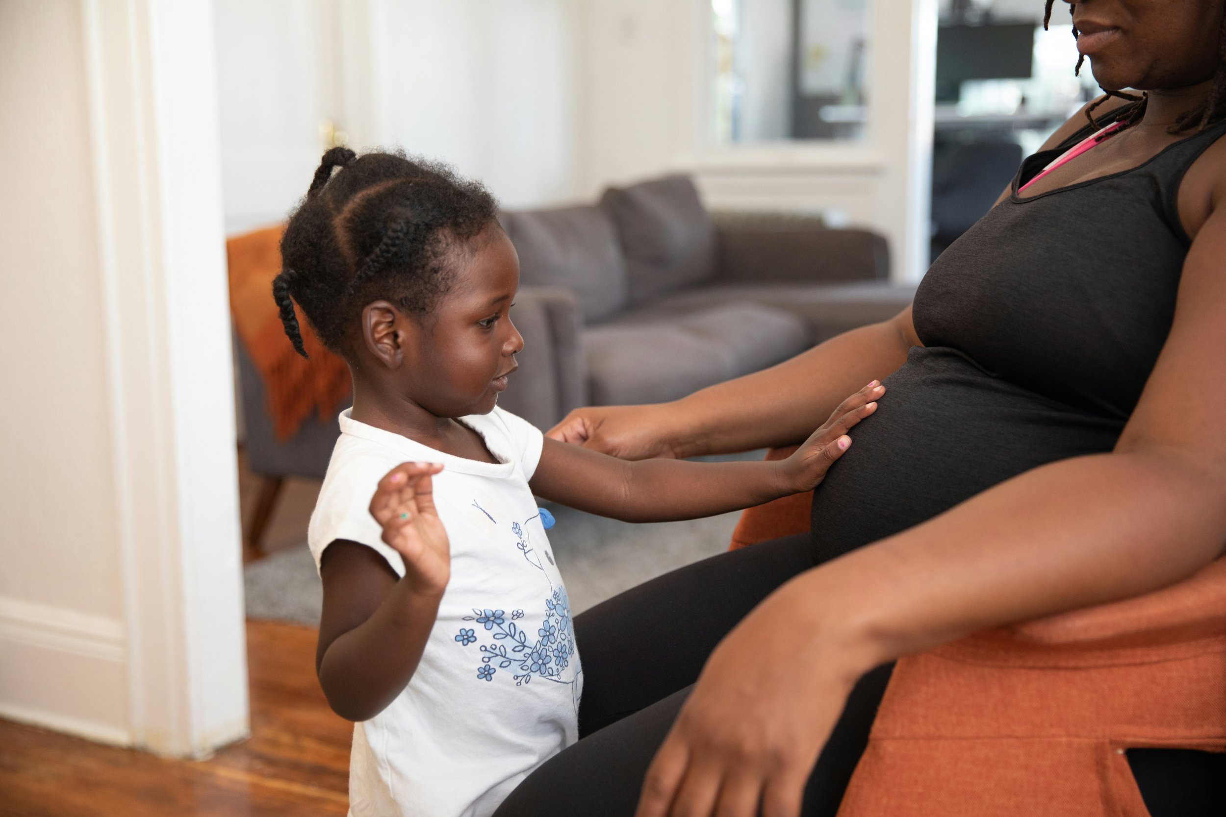 Happy, young toddler touches pregnant mother's belly as she sits in a chair