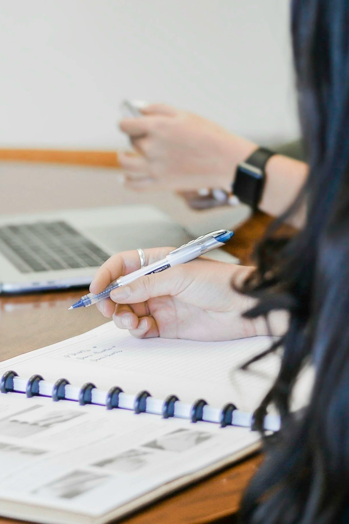 Persona tomando notas en un cuaderno en un entorno de oficina, con una computadora portátil sobre la mesa.
