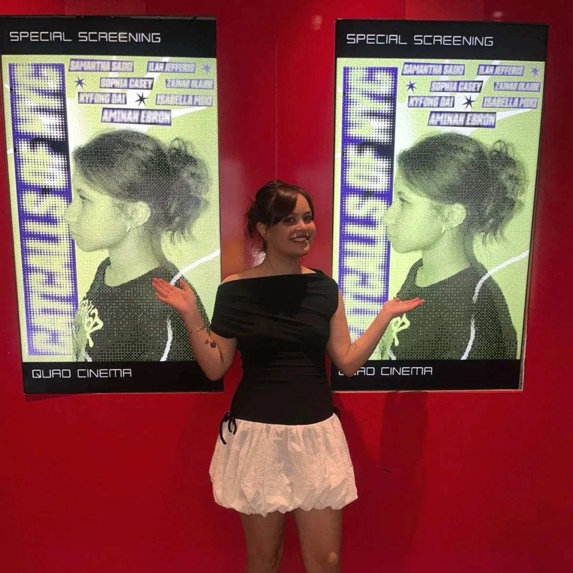 Woman smiling and gesturing with her hands in front of two large movie posters on a red wall, advertising a special screening event.