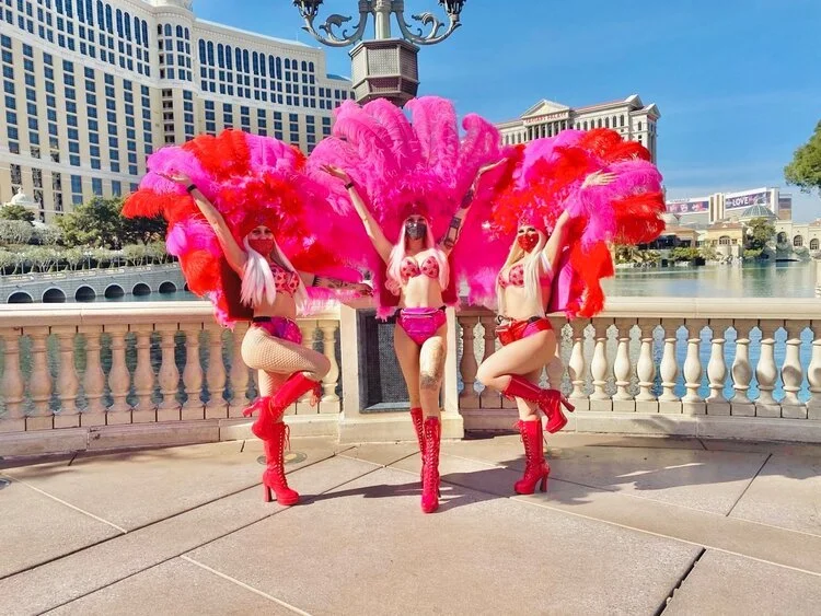 Three women dressed in colorful showgirl costumes with large pink and red feathered fans, posing outdoors on a balcony near a lake with a large hotel building in the background.