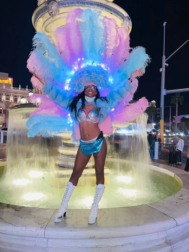 A woman in a carnival costume with large pastel feathers and lights, standing in front of a fountain at night, smiling.
