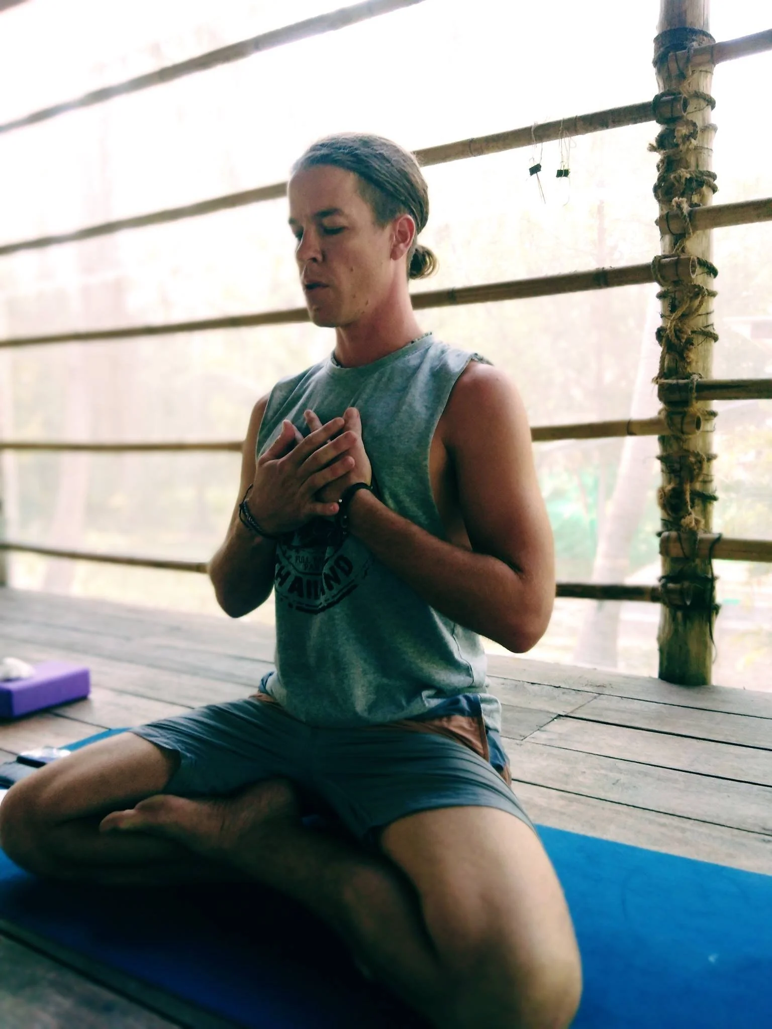 A man with long stylistic hair in a bun practicing yoga on a blue mat, sitting cross-legged with hands pressed to chest, eyes closed, in a peaceful meditation pose on a wooden platform.