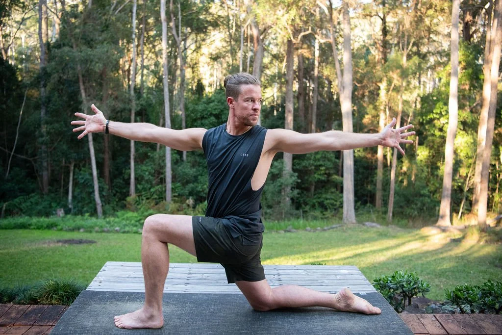 A man practicing yoga outdoors on a grassy field with a forest background during daylight, in a lunge pose with arms extended.