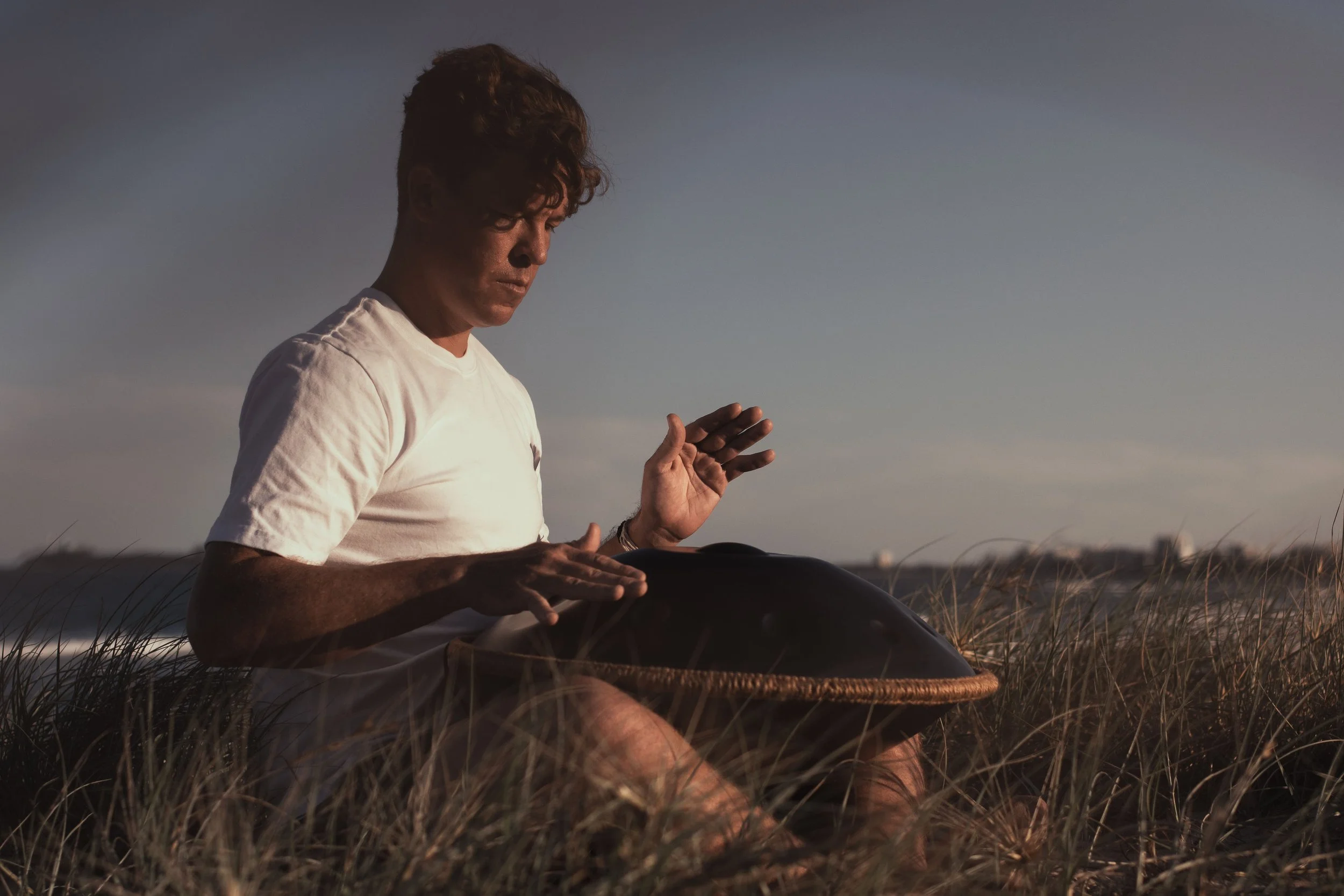 A young man in a white t-shirt sitting on the grass playing a handpan instrument during sunset.