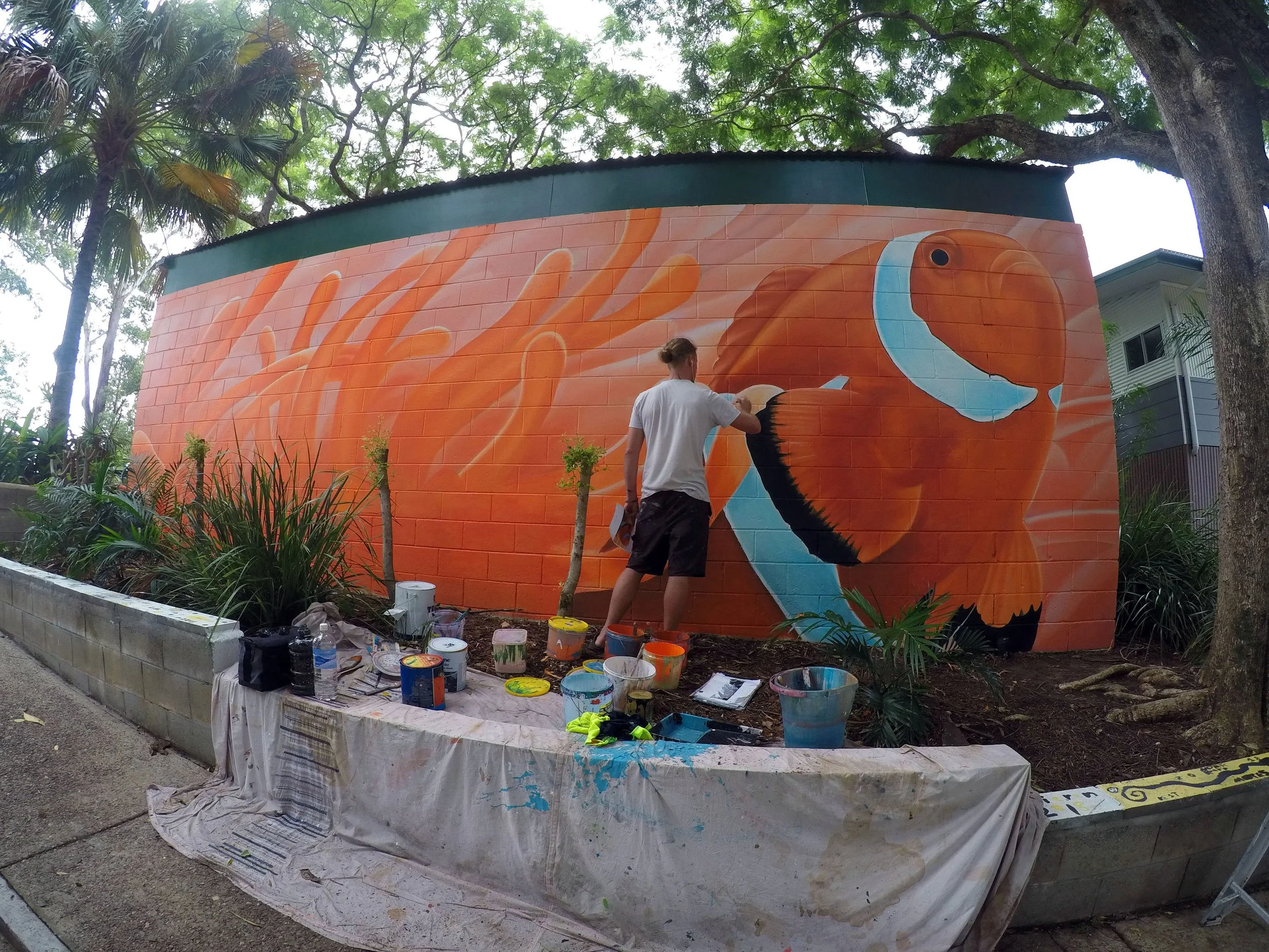 Artist painting a mural of a bald eagle on a brick wall outdoors, surrounded by trees and plants.