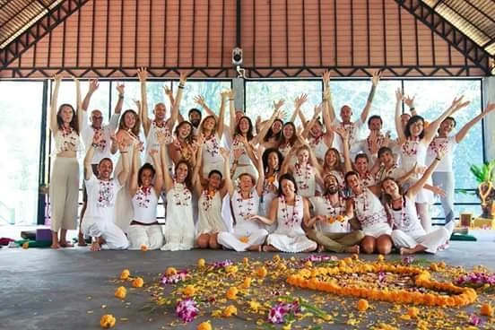 A group of women in white dresses celebrating with hands raised, standing and sitting in a studio with a decorated floor of flower petals.