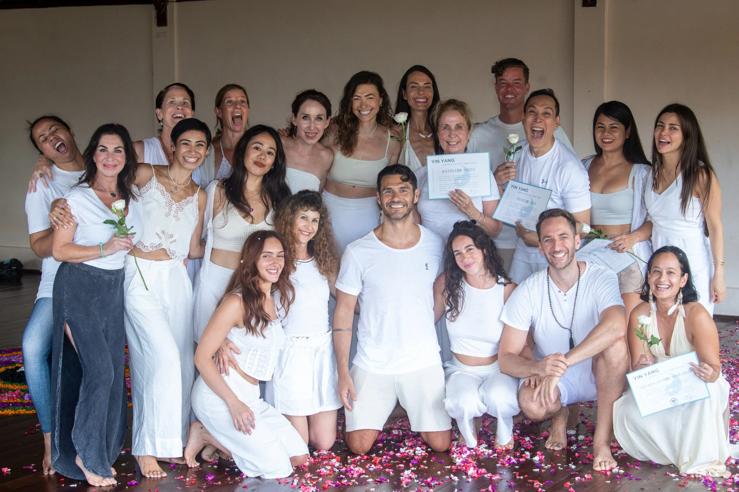 Group of people celebrating with certificates and white roses, all dressed in white, standing on a flower petal-covered floor, smiling and posing for the photo.