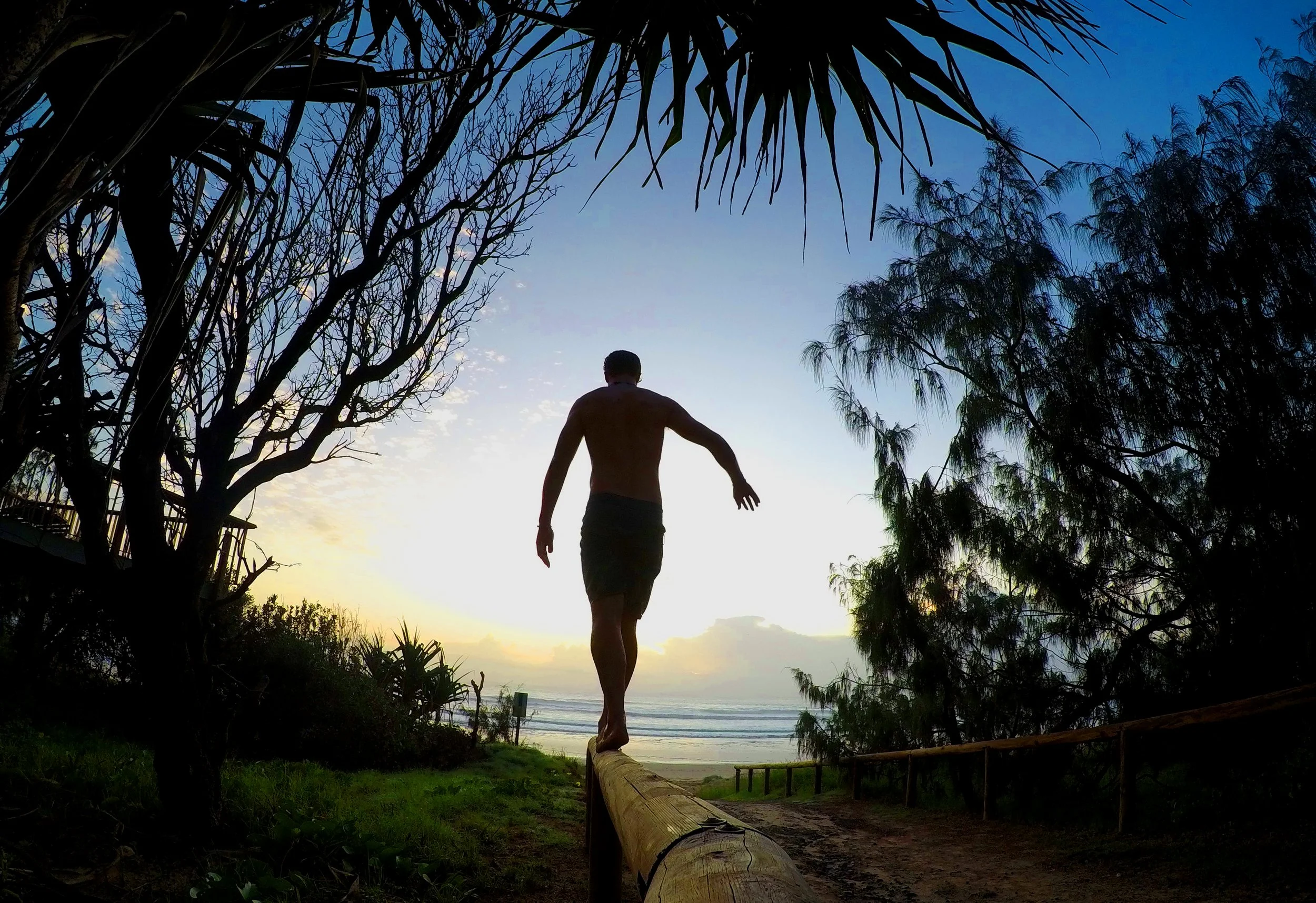 A shirtless man walking on a narrow log near the beach during sunset, framed by trees and foliage.