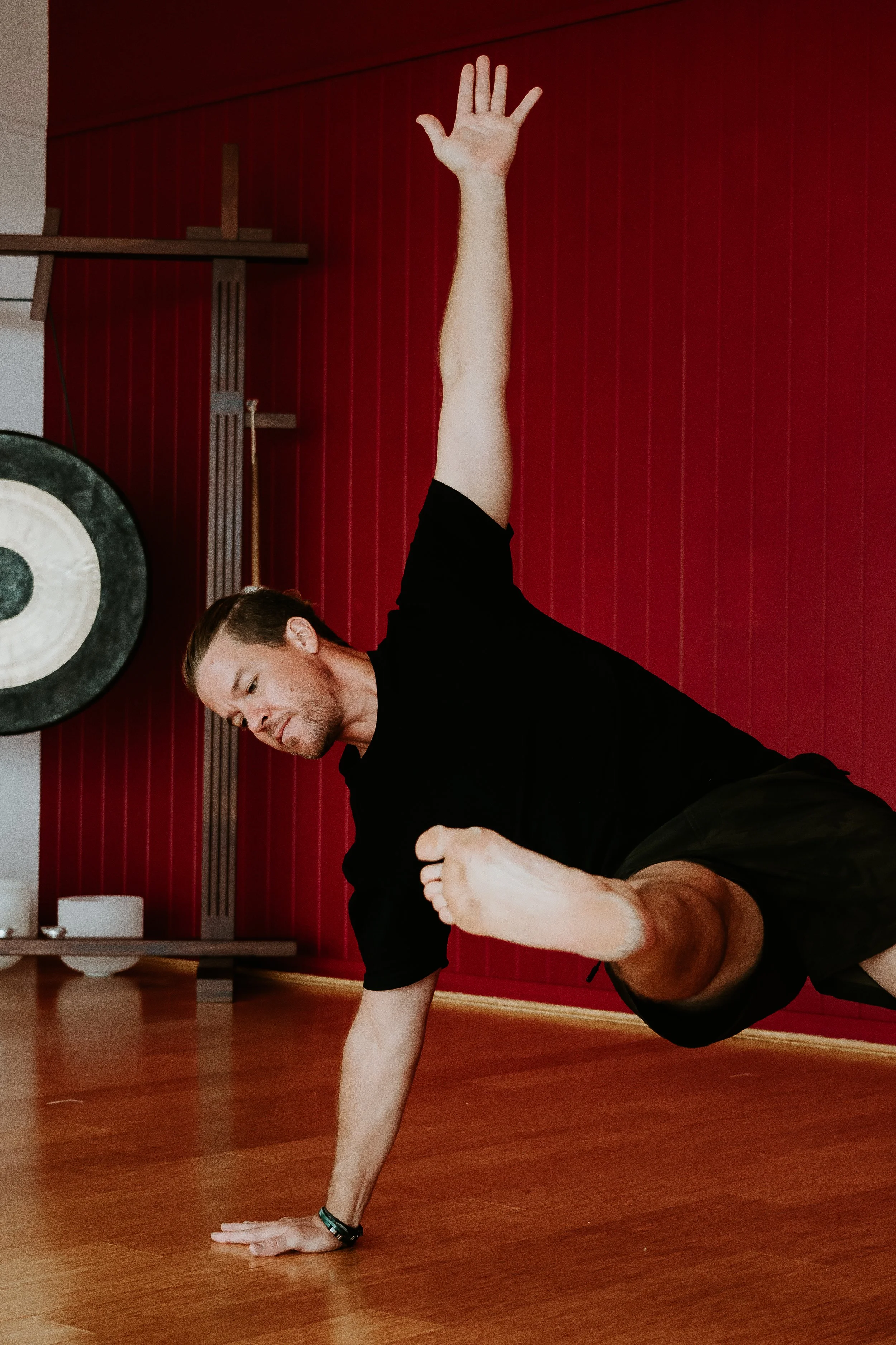 A man with long hair and a beard performing a one-arm plank exercise on a wooden floor, with a red wall and gym equipment in the background.