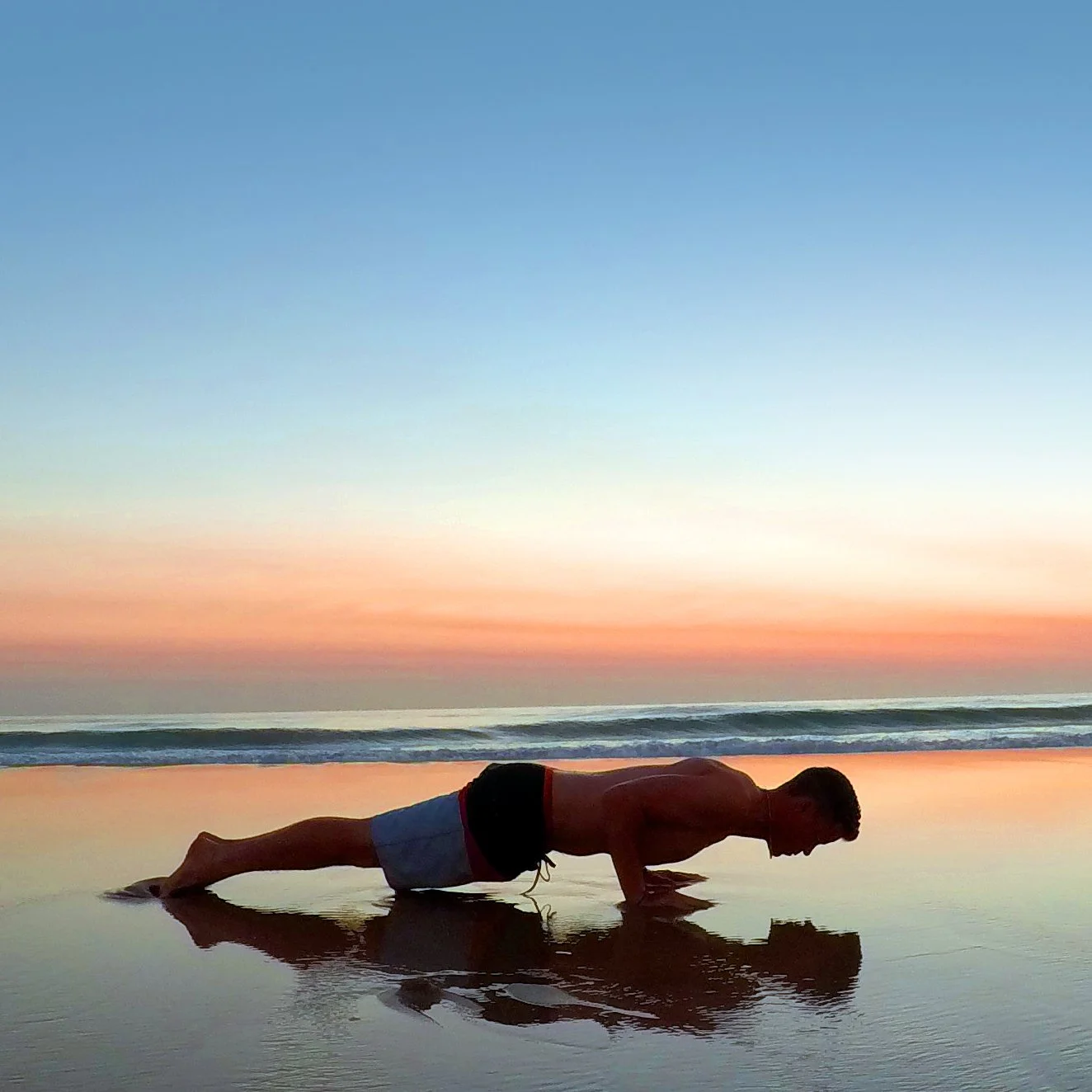 A person is doing a forearm plank on the beach during sunset or sunrise, with colorful sky and waves in the background.