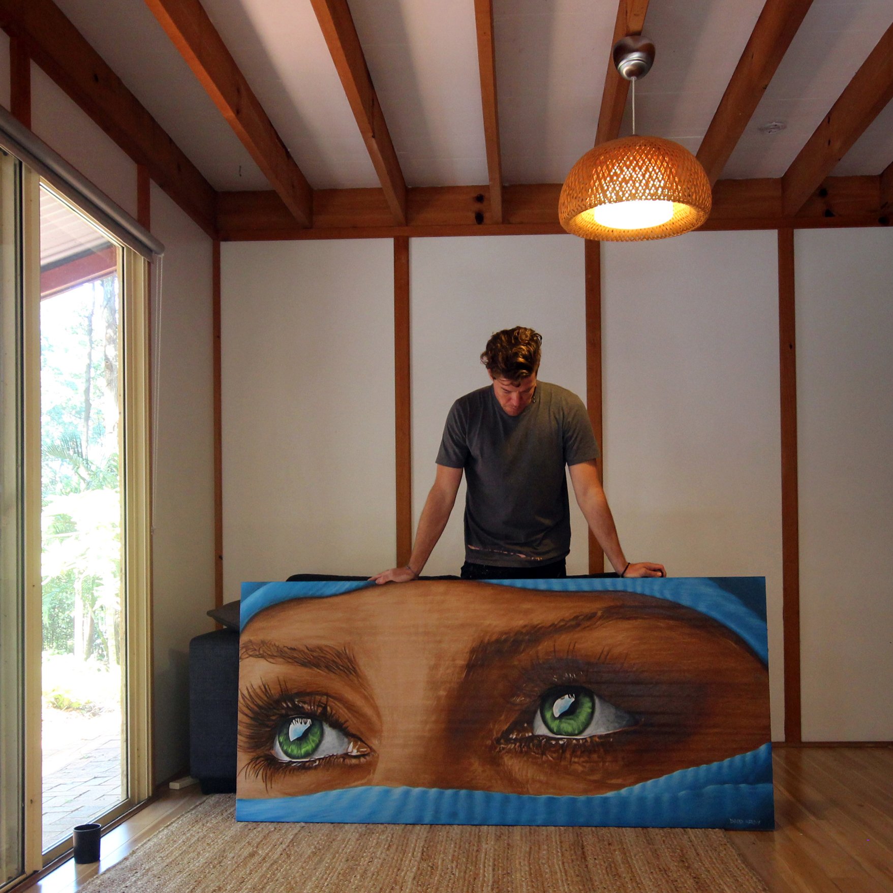 A man stands behind a large painting of realistic green eyes with eyelashes, close to a window with a sliding door, in a room with wooden ceiling beams and a wicker light fixture.