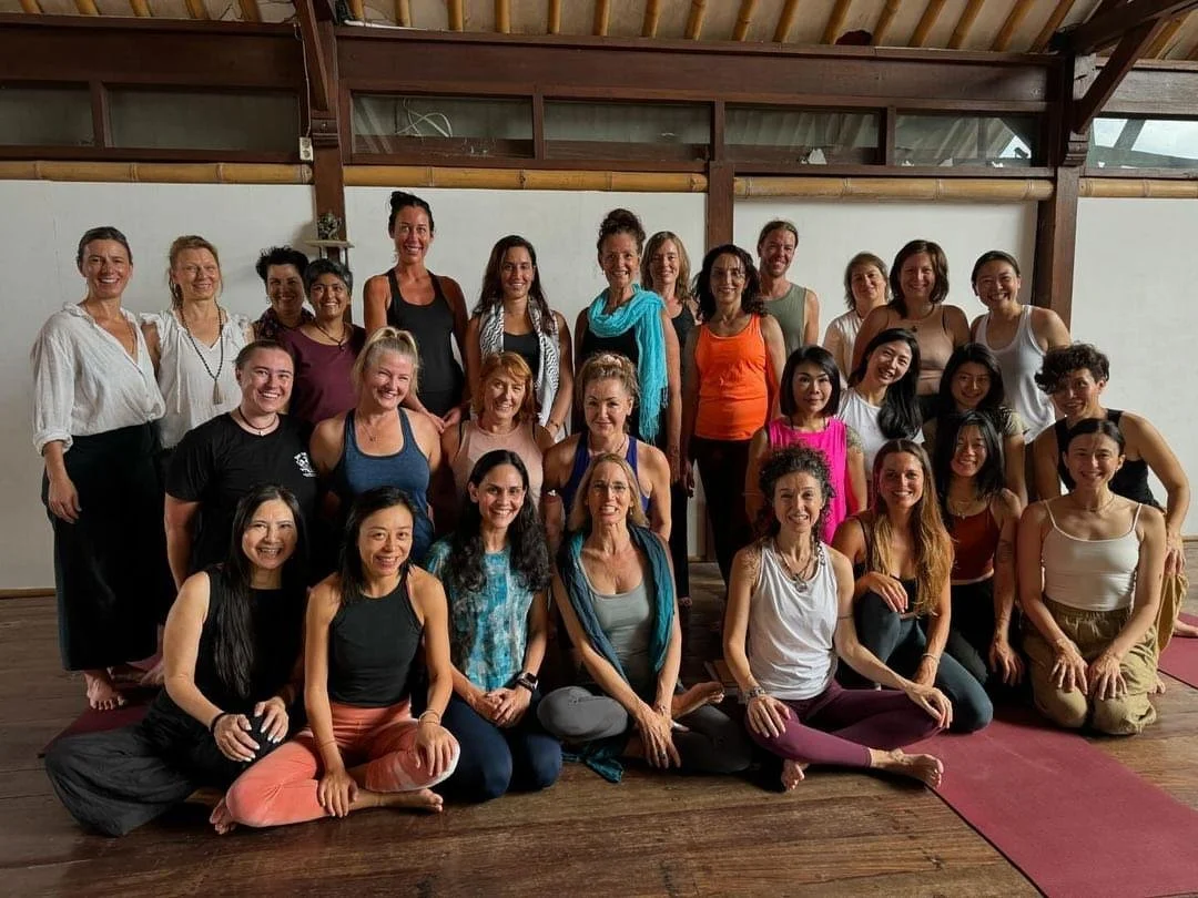 Group of women gathered in a yoga or wellness studio, posing for a group photo, some sitting and others standing, with yoga mats on the floor.