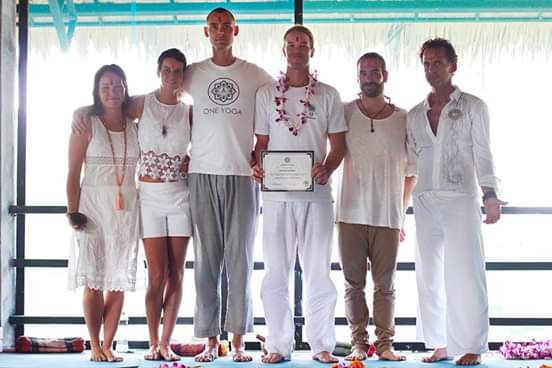 Group of six people standing on a stage, one holding a certificate, in front of a large window with light curtains.