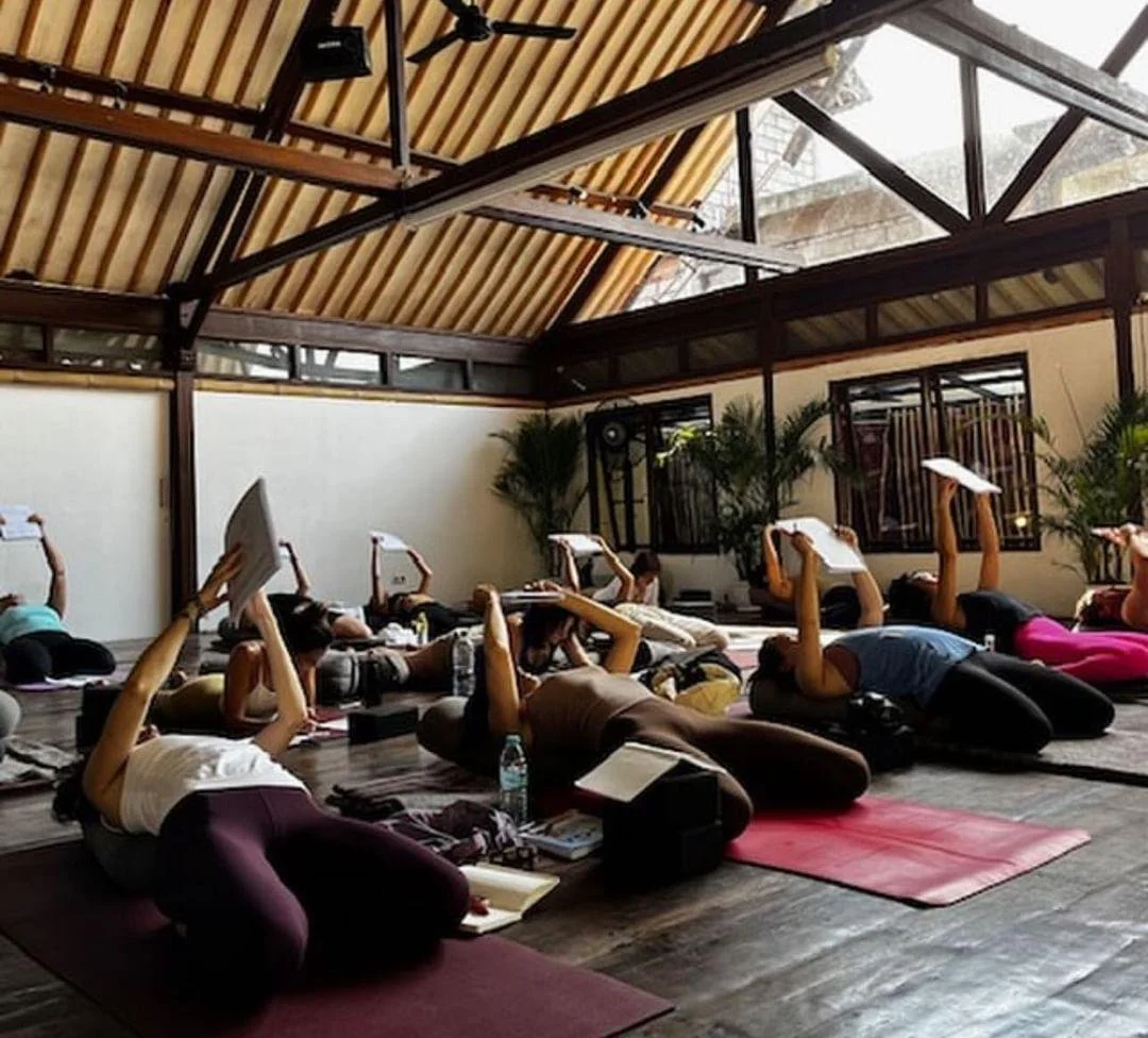 People lying on yoga mats in a spacious, well-lit yoga studio, participating in a class while holding notebooks.
