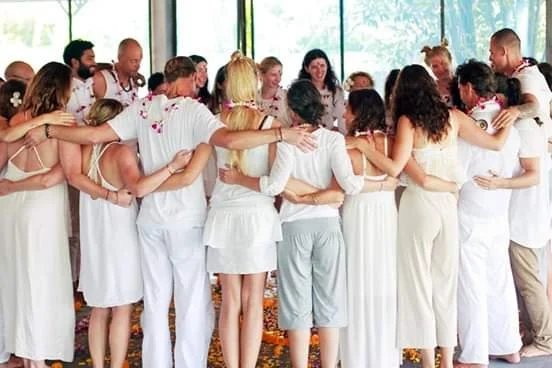 A diverse group of people dressed in white, standing in a circle with their arms around each other indoors, engaged in a supportive group activity or celebration.