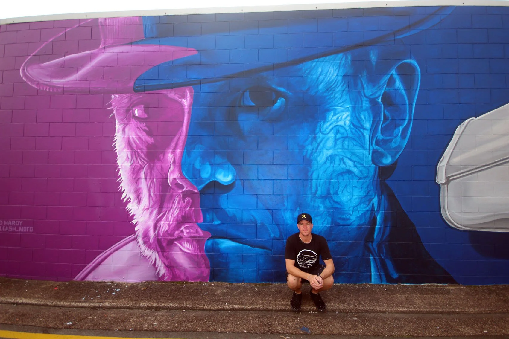 A man squatting in front of a colorful street mural of two faces, one in pink with a police hat and beard, and the other in blue with glasses, on a brick wall.