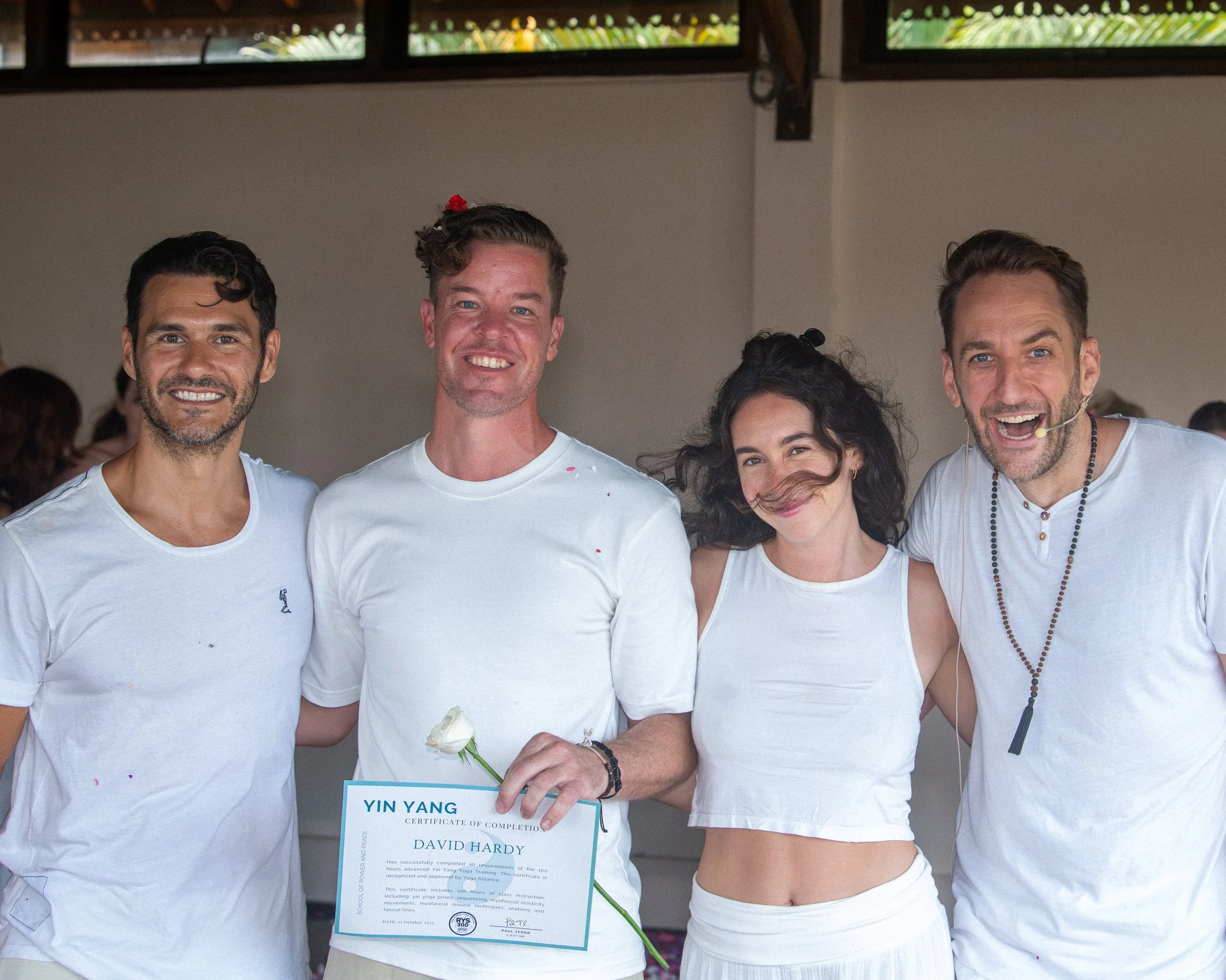 Group of four people at a celebration, with one person holding a certificate and a flower, all smiling and dressed in white.