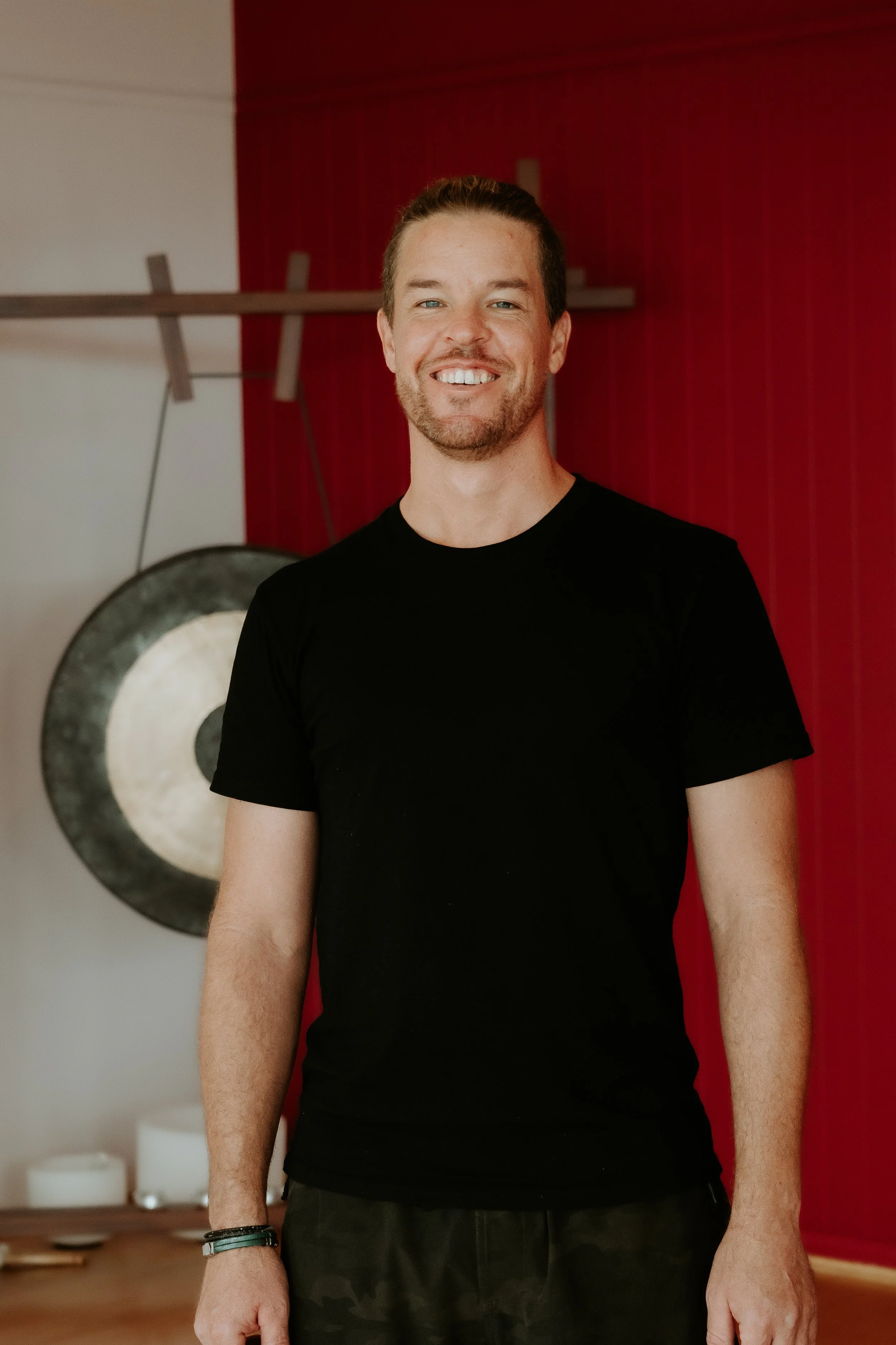 A smiling man in a black t-shirt standing indoors in front of a red wall with a gong and some white items visible in the background.